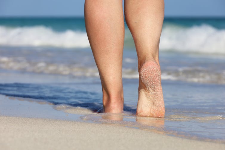 Frau mit nackten Beinen und Futter im seichten Meerwasser beim Spaziergang am Sandstrand, weiße Wellen im Hintergrund
