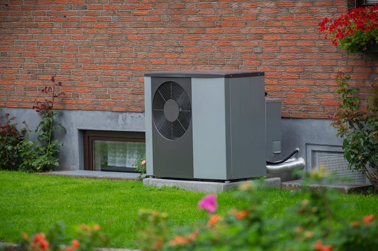 Graue Luftwärmepumpeneinheit an der Fassade eines alten roten Backsteinhauses, grünes Gras und Blumen im Vordergrund