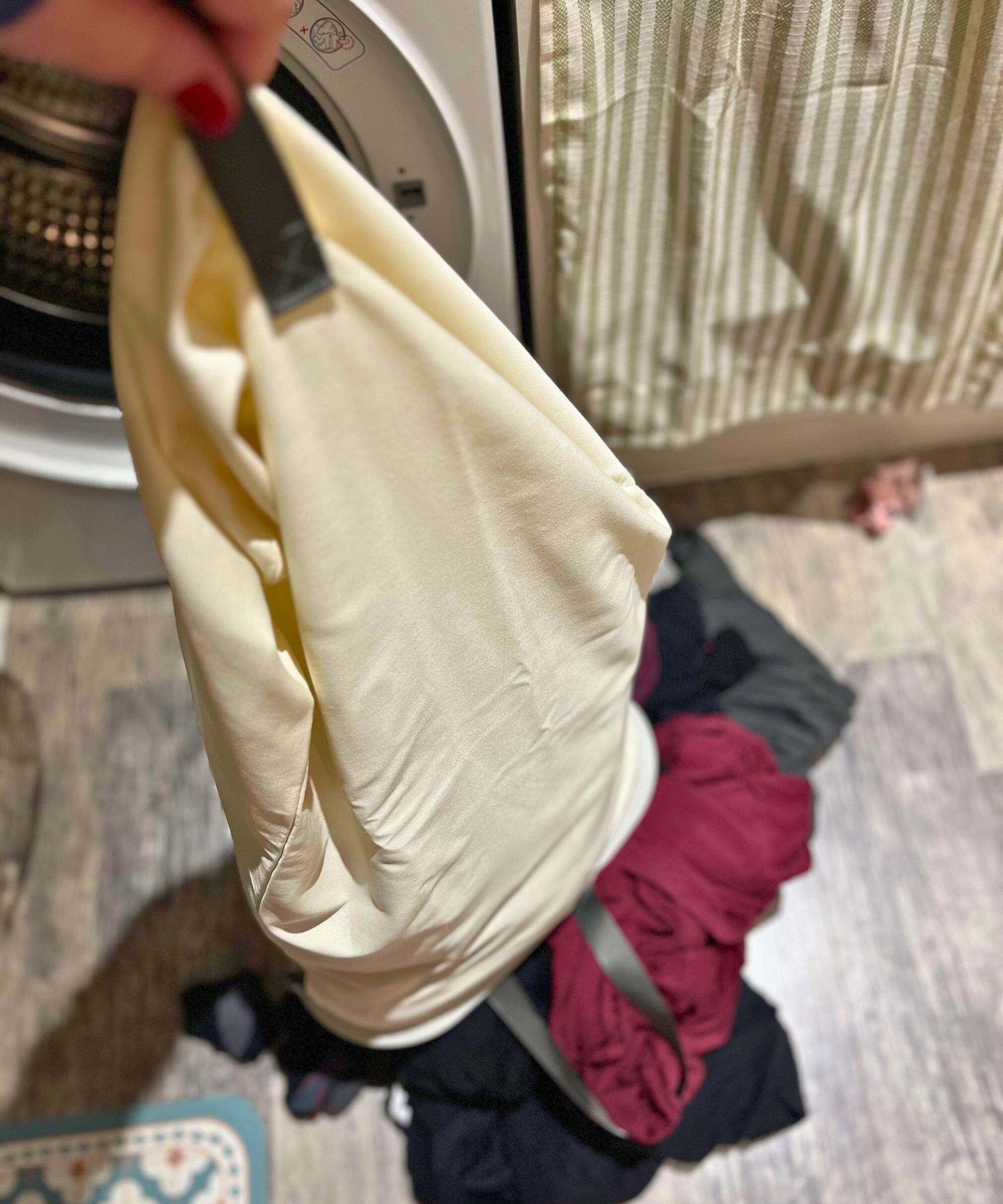 Ein cremefarbener Leinensack mit schwarzen Henkeln voller Wäsche wird auf den grauen Linoleum-Küchenboden neben der Waschmaschine geleert