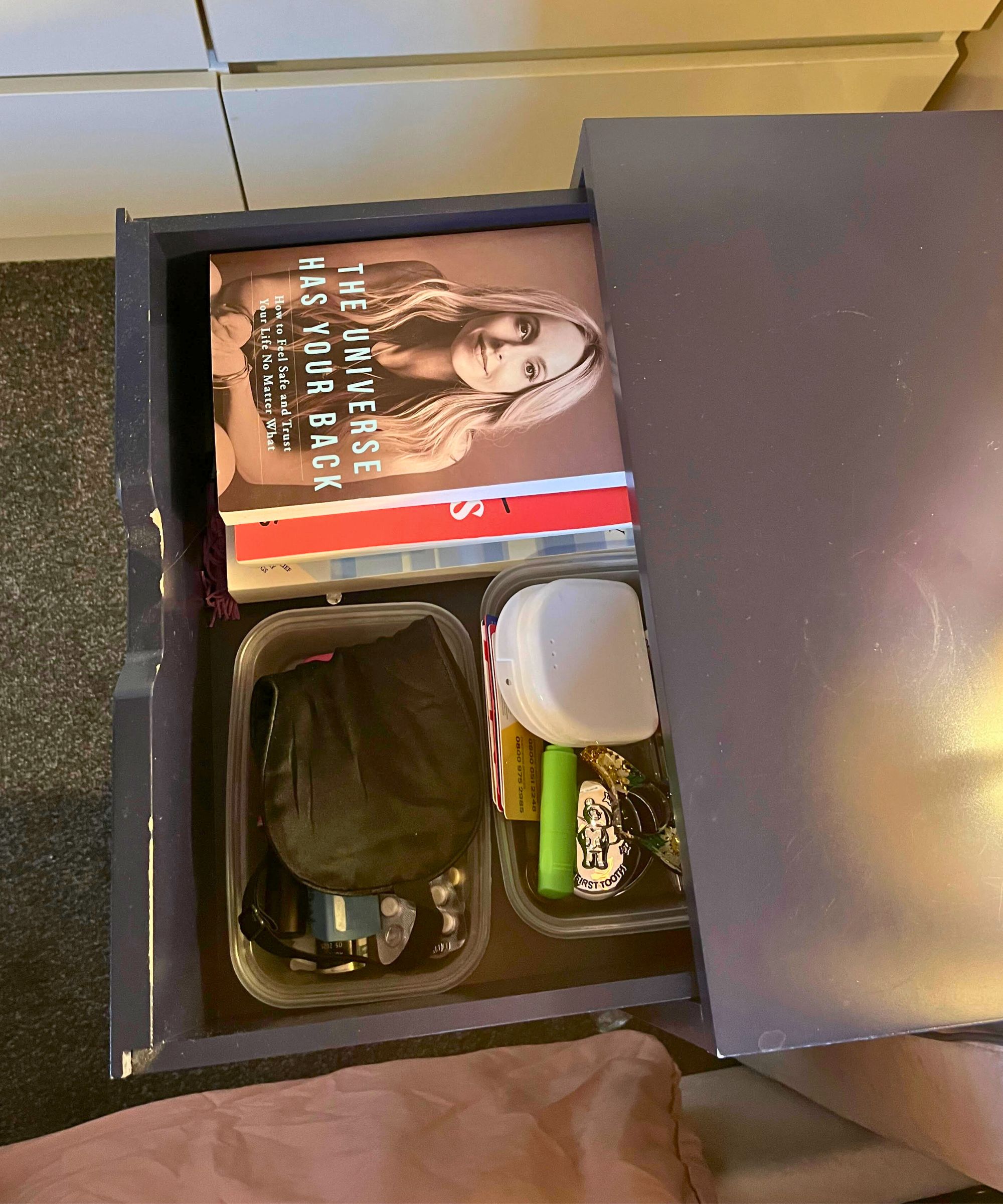 Nachttischschublade danach, sehr ordentlich, mit einem Stapel Bücher auf einer Seite und ein paar Plastikbehältern mit einer ordentlichen Auswahl an Gegenständen, darunter Schlafmaske und Medikamente