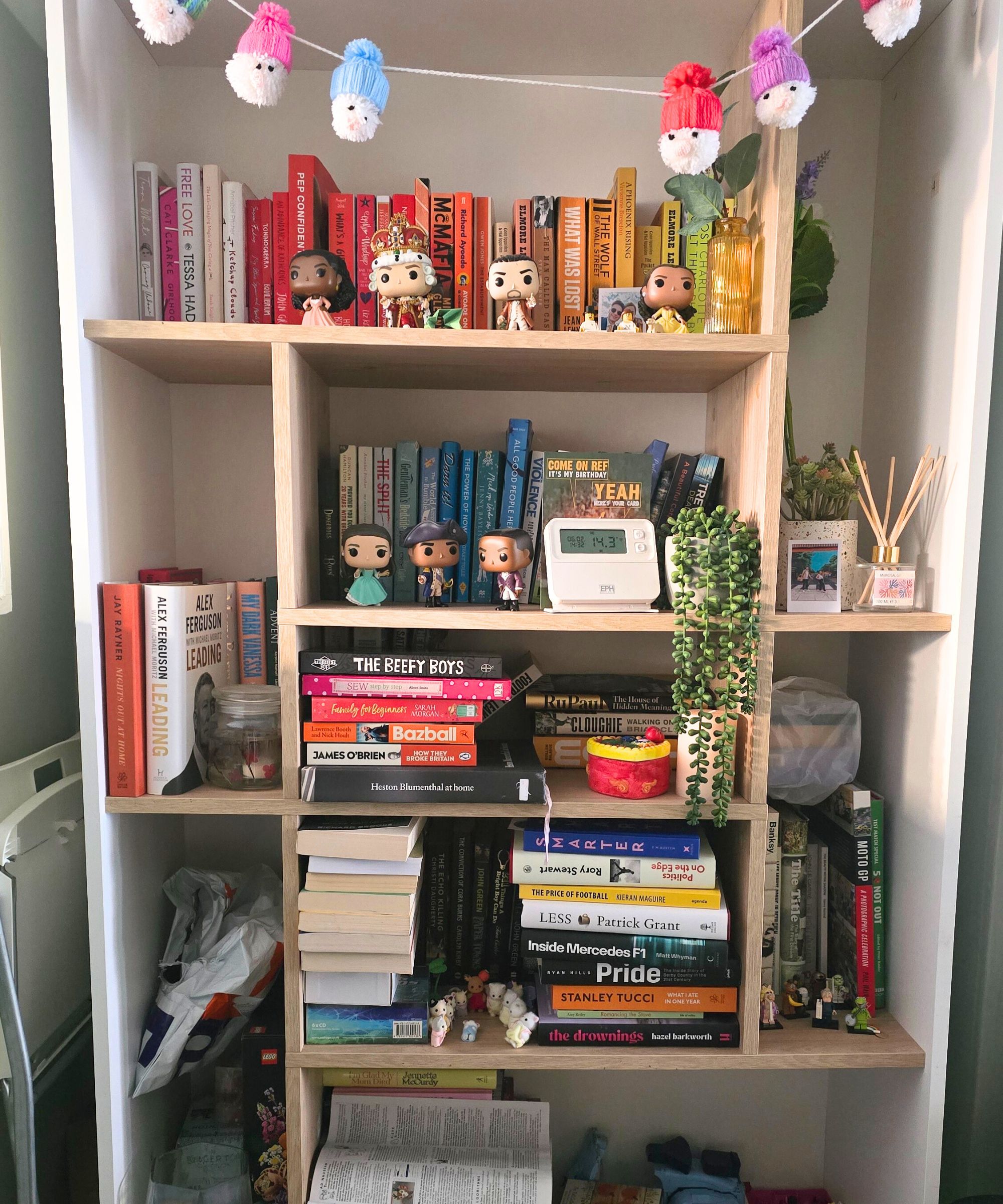 Ein weißes Bücherregal mit Santa -Dekorationen, die über die Spitze hängen, und farbenfrohe Bücher, Kunstpflanzen und Funko pops darauf