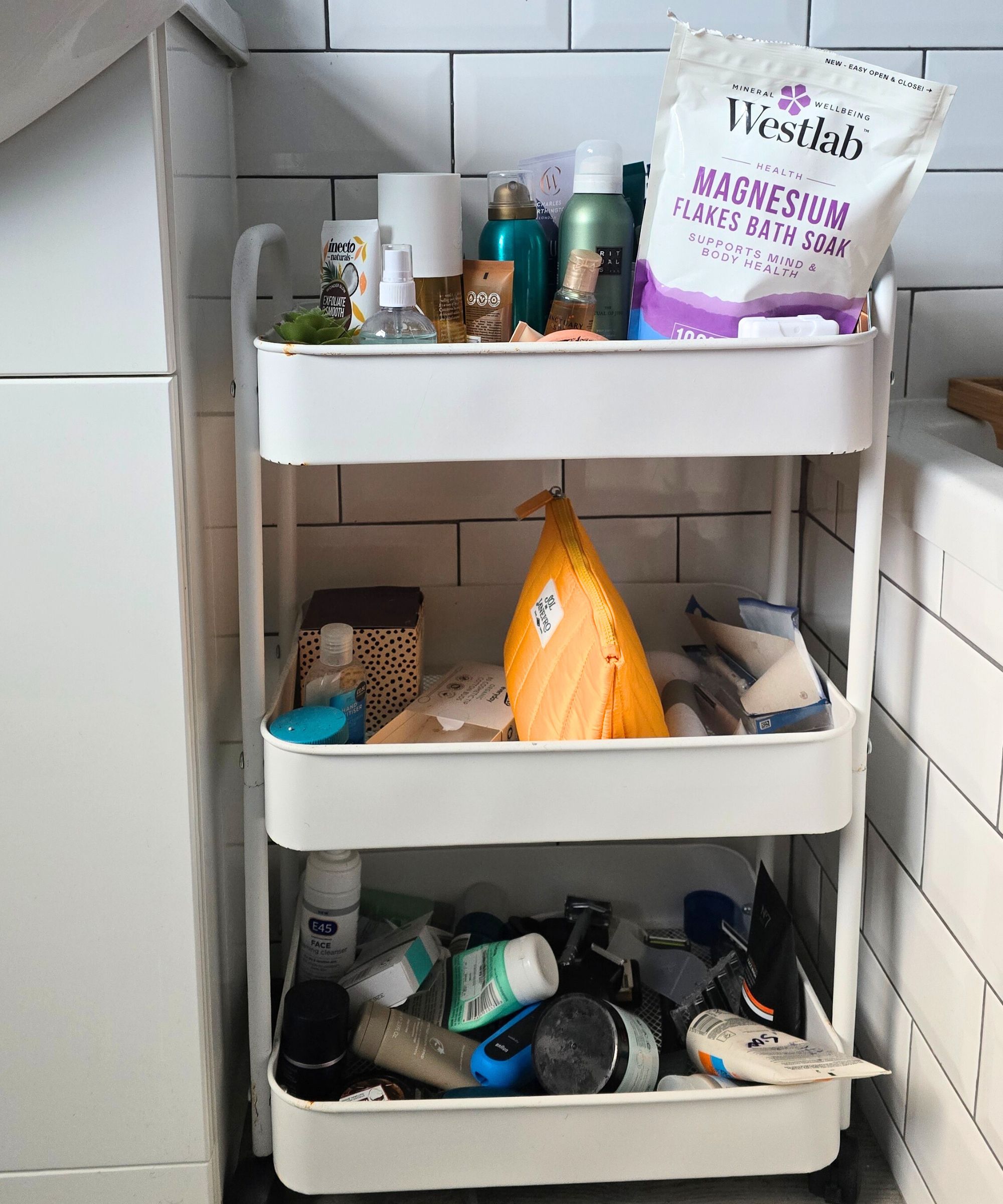 Ein überfüllter dreistufiger weißer Aufbewahrungswagen mit farbenfrohen Toilettenartikeln und weiße Fliesen dahinter