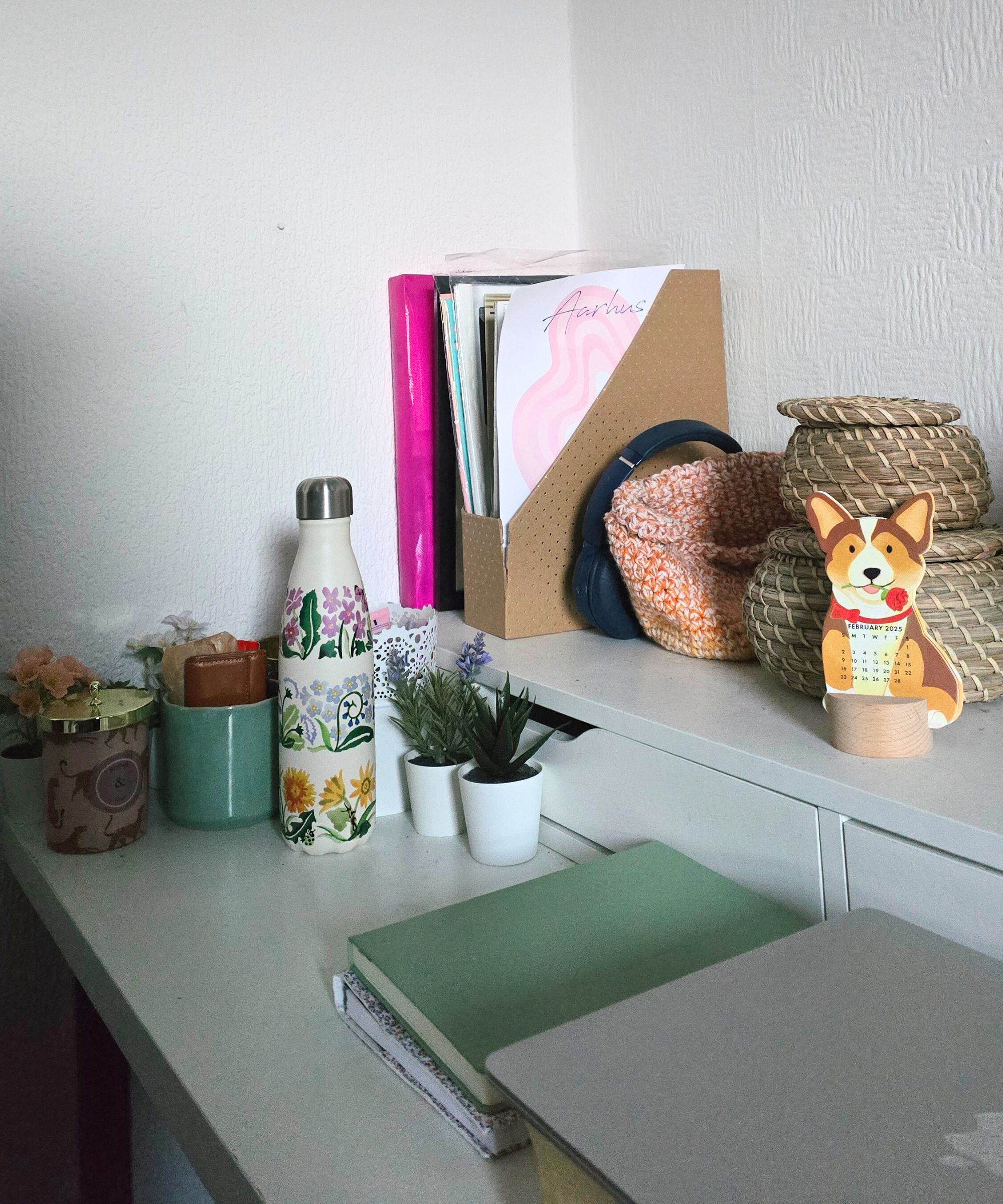 Ein weißer zweistufiger Schreibtisch mit kreisförmigen gewebten Wannen, einem Magazinfiler, Kerzen, einer Wasserflasche, einem grünen Notizblock und einem silbernen Laptop darüber hinaus
