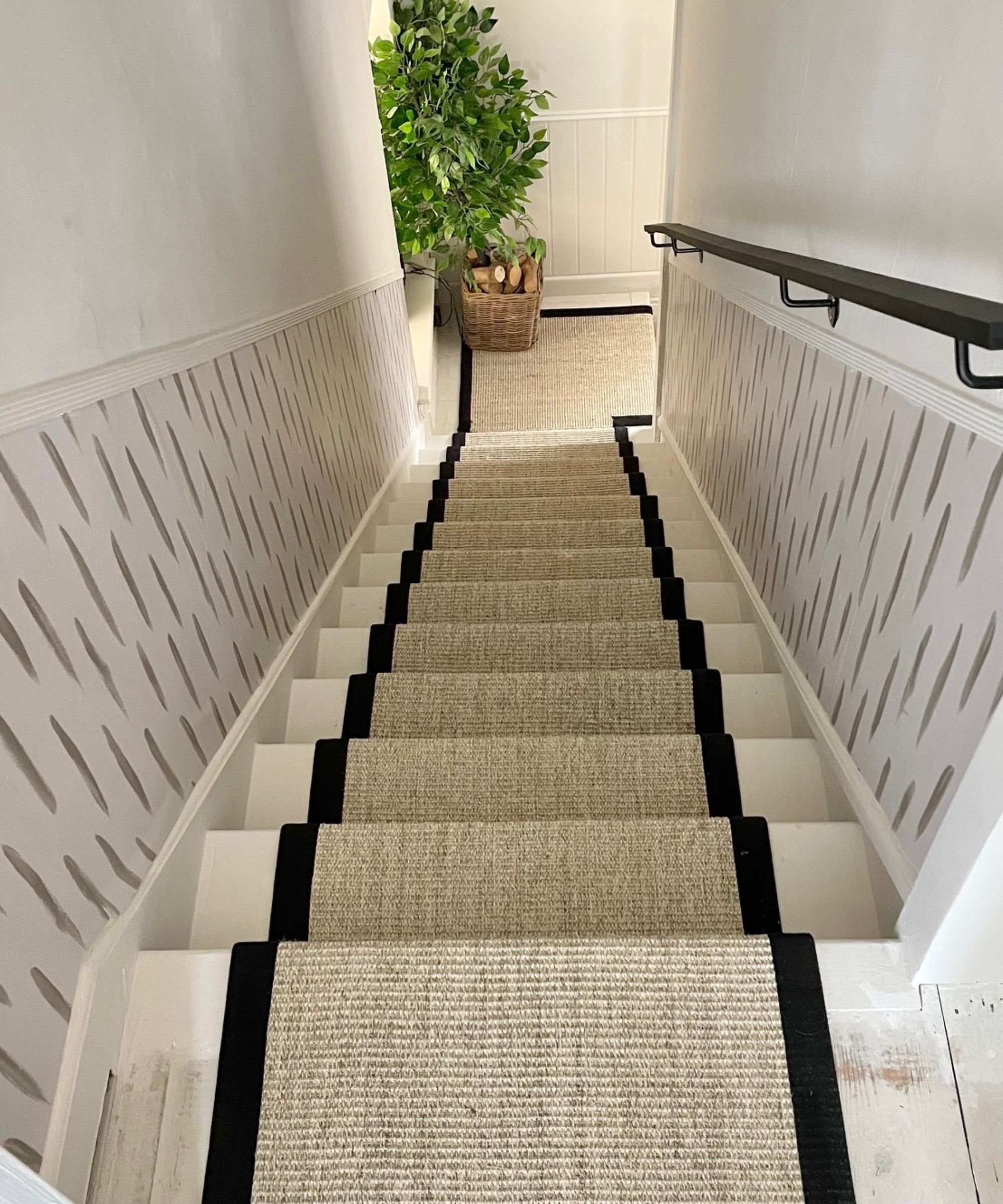 Eine Abwärtsaussicht auf einen beige gewebten Treppenläufer mit schwarzer Kante auf einer weißen Treppe mit einem schwarzen Handlauf und einer großen Pflanze unten