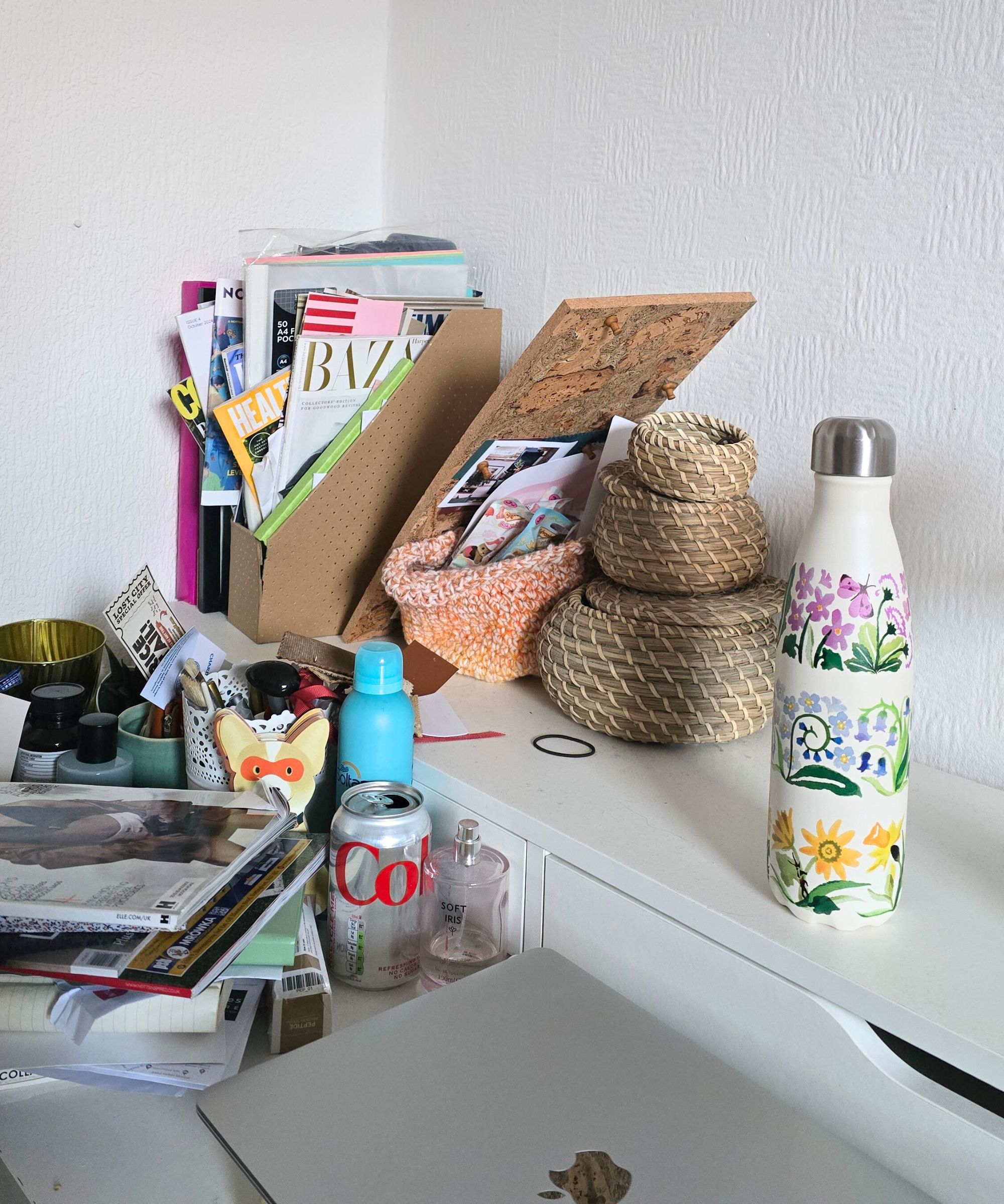 Ein weißer Schreibtisch mit einer Wasserflasche, gewebten kreisförmigen Kisten, einem vollständigen Magazinfiler, einem Haufen Magazine und einem silbernen Laptop darüber