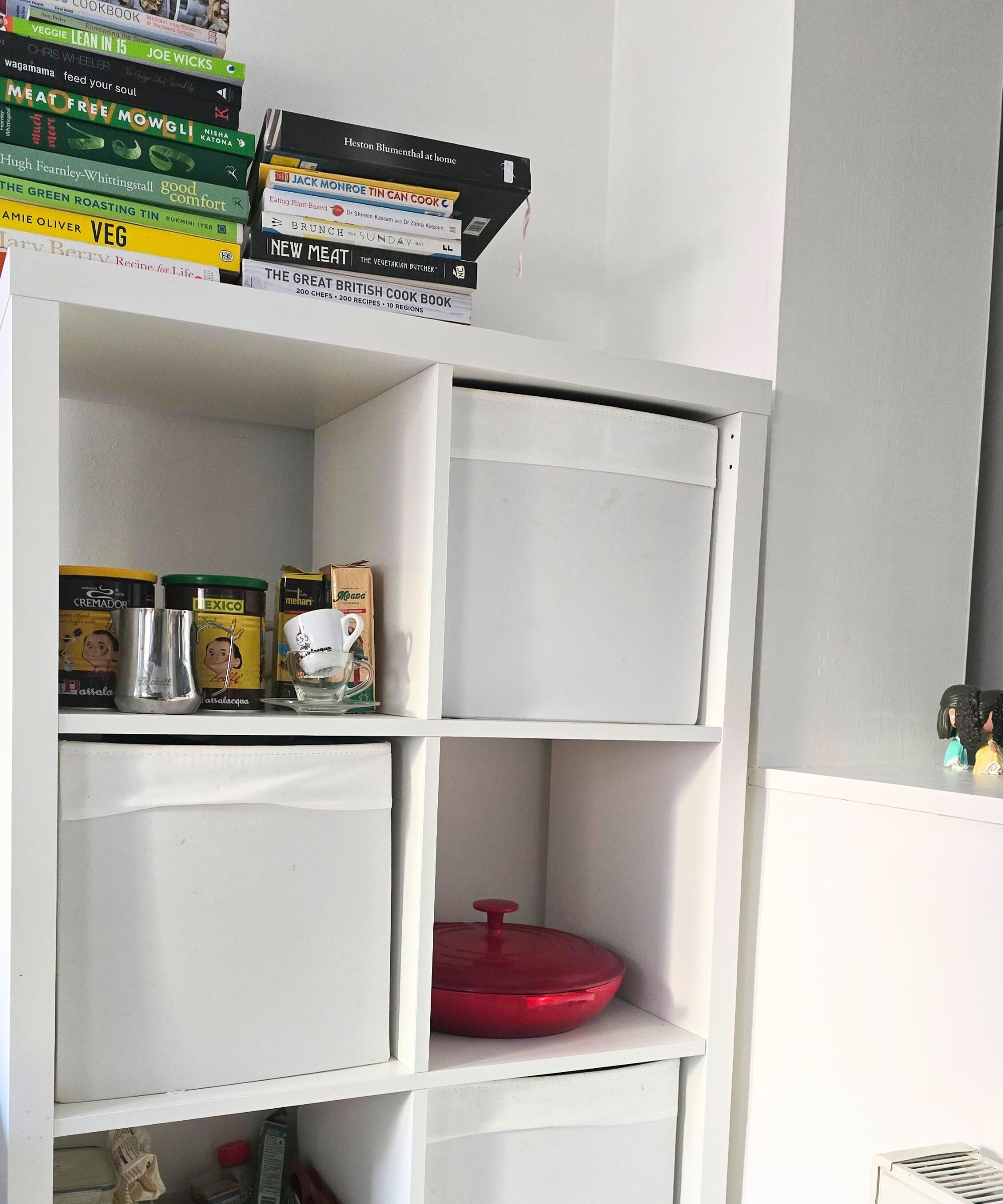 Eine weiße Küchenlagereinheit mit farbenfrohen Büchern darüber und Kaffeezubehör, eine rote Küchenpfanne und weiße Aufbewahrungsboxen darin