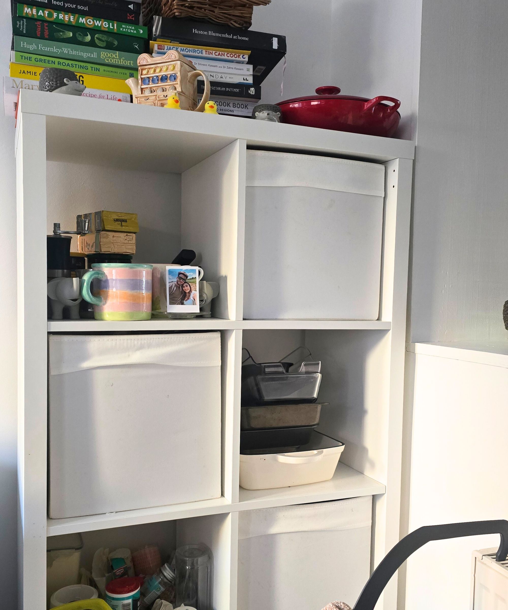 Eine weiße Küchenlagereinheit mit Büchern, eine Teekanne und eine rote Küchenpfanne darüber, Tassen, Schalen, Backzutaten und weiße Lagerwürfel darin