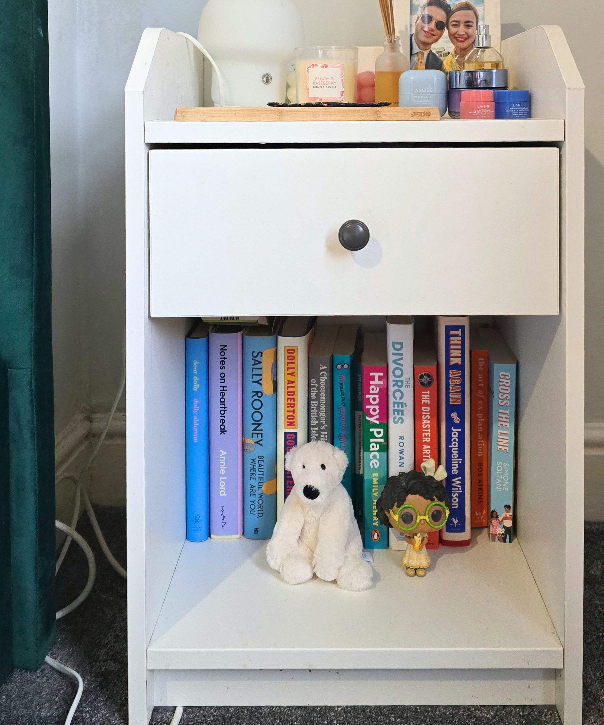 Ein weißer Nachttisch mit farbenfrohen Büchern, ein Polarbärenspielzeug und ein Funko -Pop im unteren Regal, Make -up -Wannen, eine Lampe und ein Fotorahmen im oberen Regal