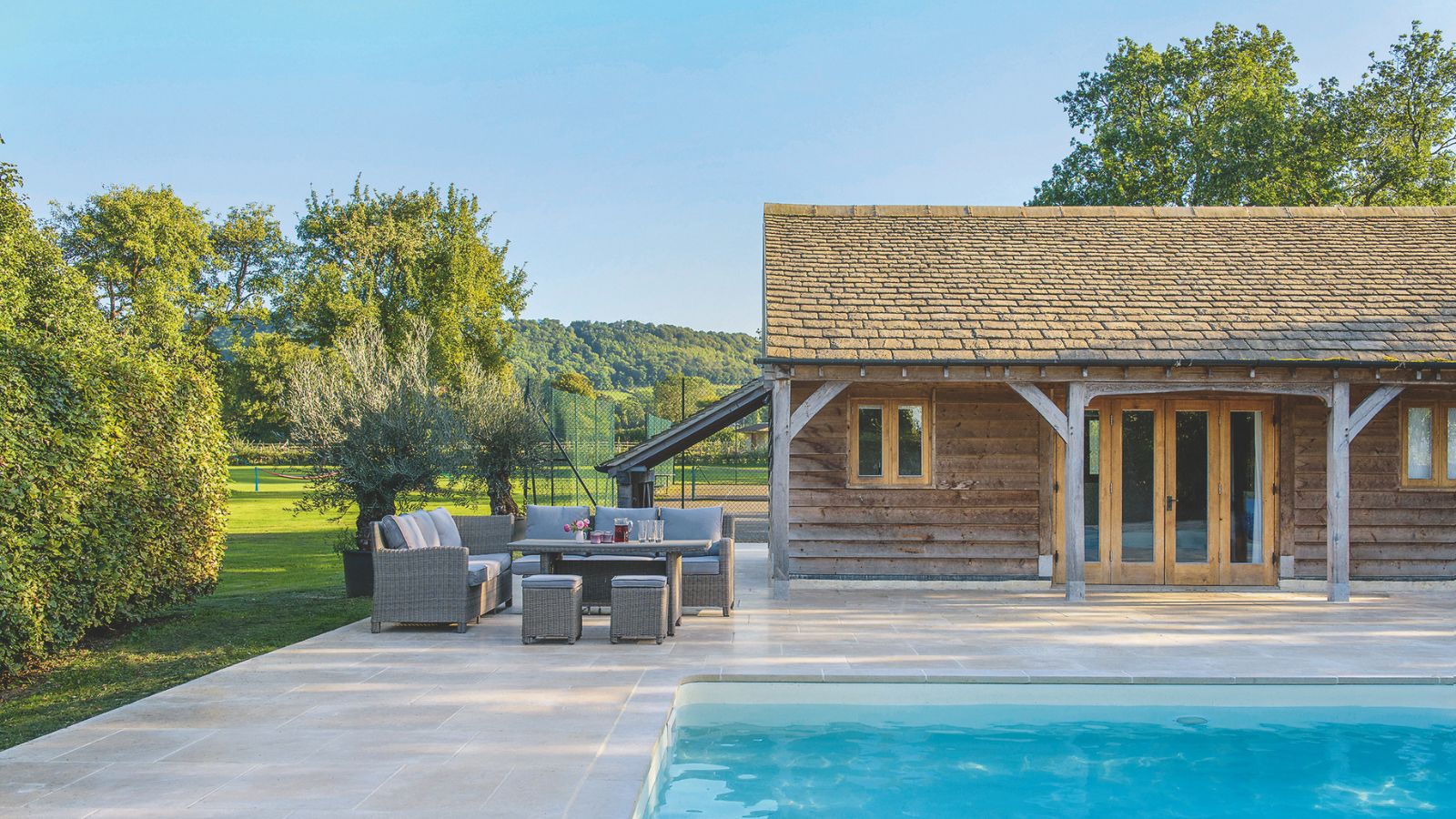 Großer sehr sauberer und gut gepflegter, gepflasterter Bereich im Freien, der Poolhaus, Essmöbel im Freien und die Rand eines Pools im Vordergrund zeigt