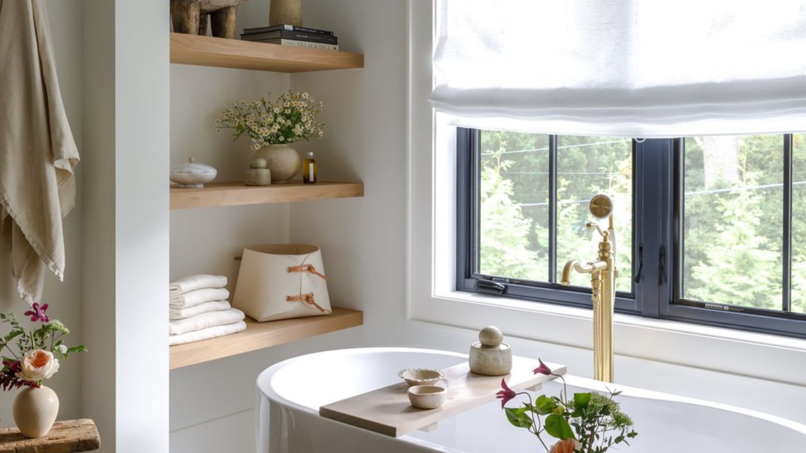 Bright white bathroom corner with deep bathtub, wooden shelves with neatly organized towels and bath products, and hanging towels. there are multiple vases of flowers in shot and a linen window treatment
