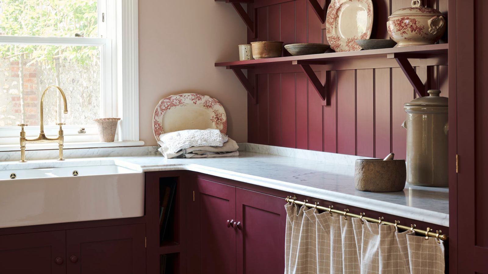 Burgundy cottage laundry room with white counters, gold rail across the cabinet with a curtain instead of a door. There are peach and cream china plates on display on a shelf