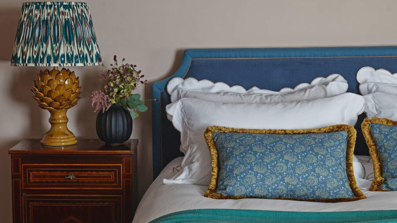 White and blue pillows on a bed with a blue headboard against cream walls; to the left, a lamp and a vase on a wooden nightstand.