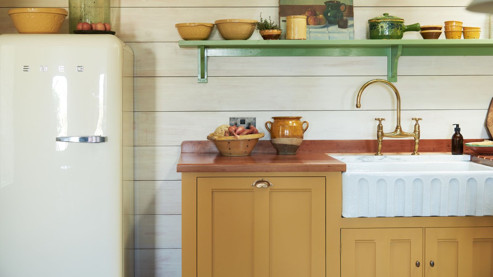 Nahaufnahme einer geriffelten Unterbauspüle mit roten Arbeitsflächen und grünen Regalen über der Spüle. Links ist ein weißer SMEG-Kühlschrank zu sehen