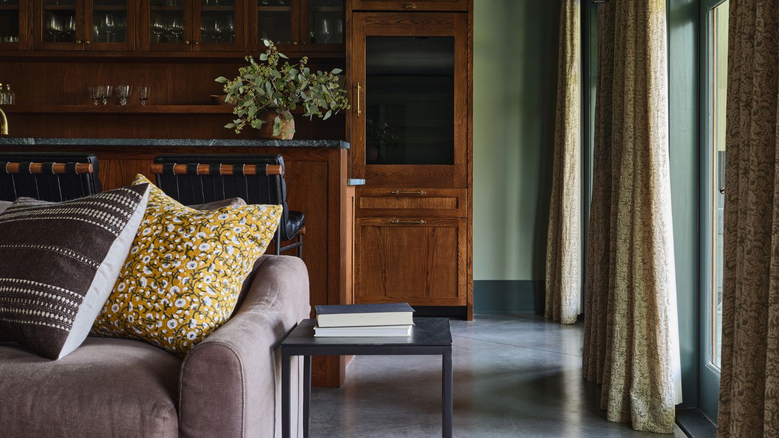 Rustic living room area, partially pictured is a velvet sofa with yellow and black cushions, a small side table, and in long floor-to-ceiling curtains. In the background is a dining area with high stools and wooden furnishings.