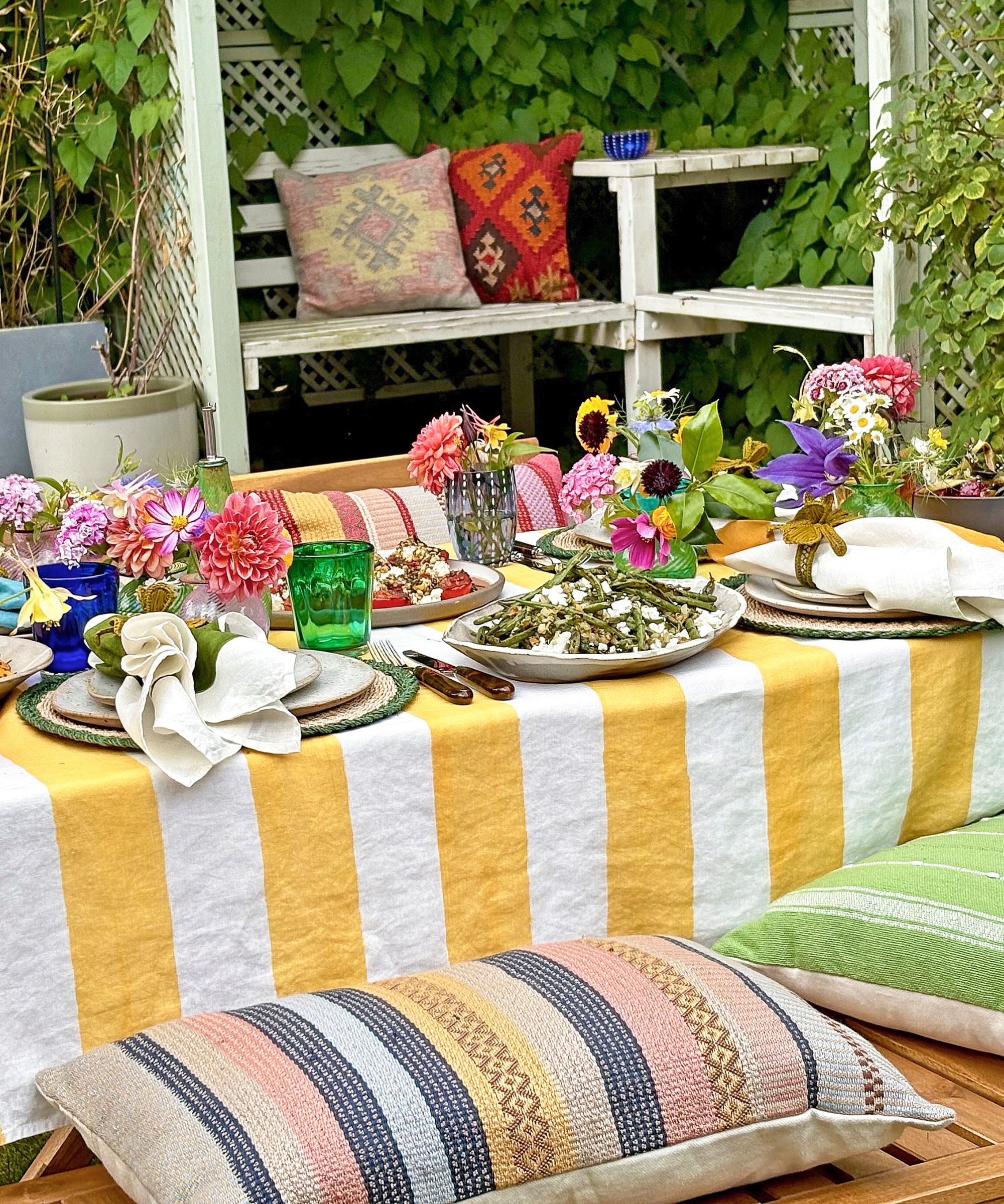 Buntes Sommertisch im Freien mit Blumen, Frucht- und Salatenplatten, einer gelben und weißen gestreiften Tischdecke und farbigen Glaswaren mit Kissen auf einer Bank
