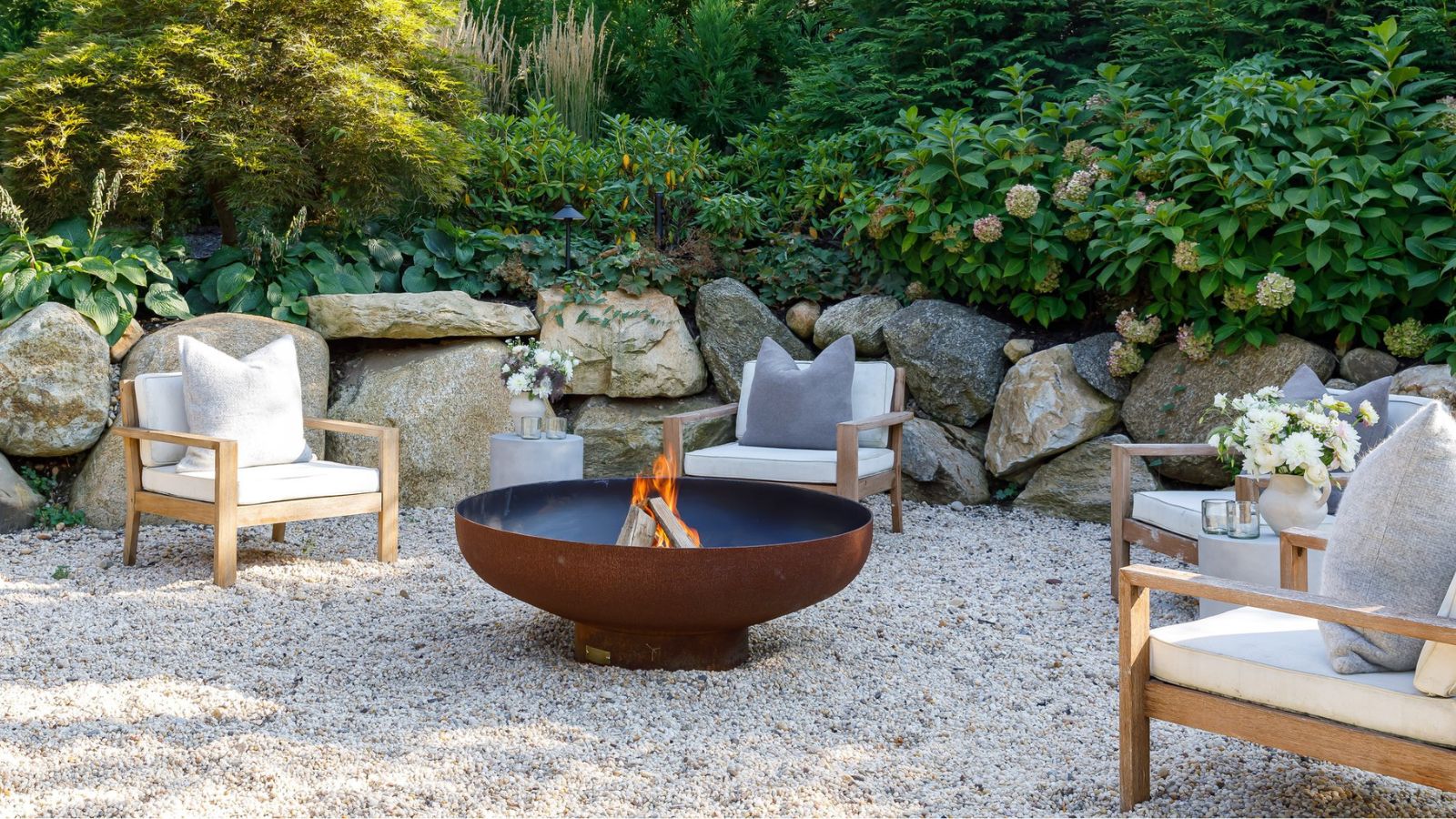 Four outdoor wooden chairs with deep white cushions and throw pillows, spaced out around a low-profile round metal fire pit. The floor is pebbled, there is a stacked rock border and everygreen shrubs in the background