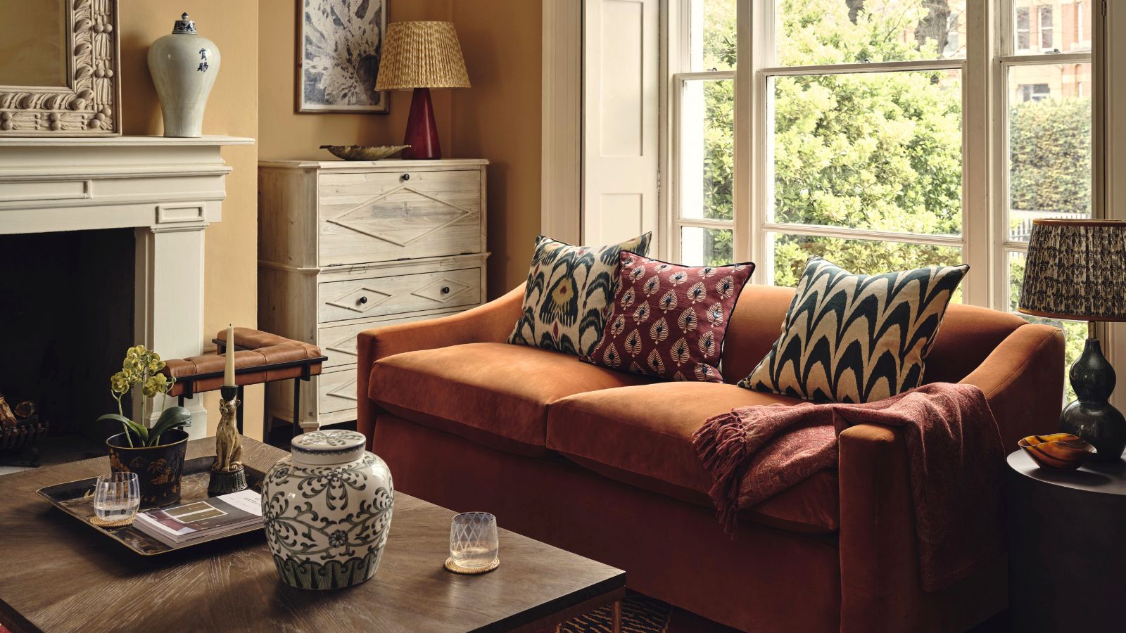 A large orange fabric sofa in a warm toned living room, in front of a large window. A large wooden coffee table in front of it.