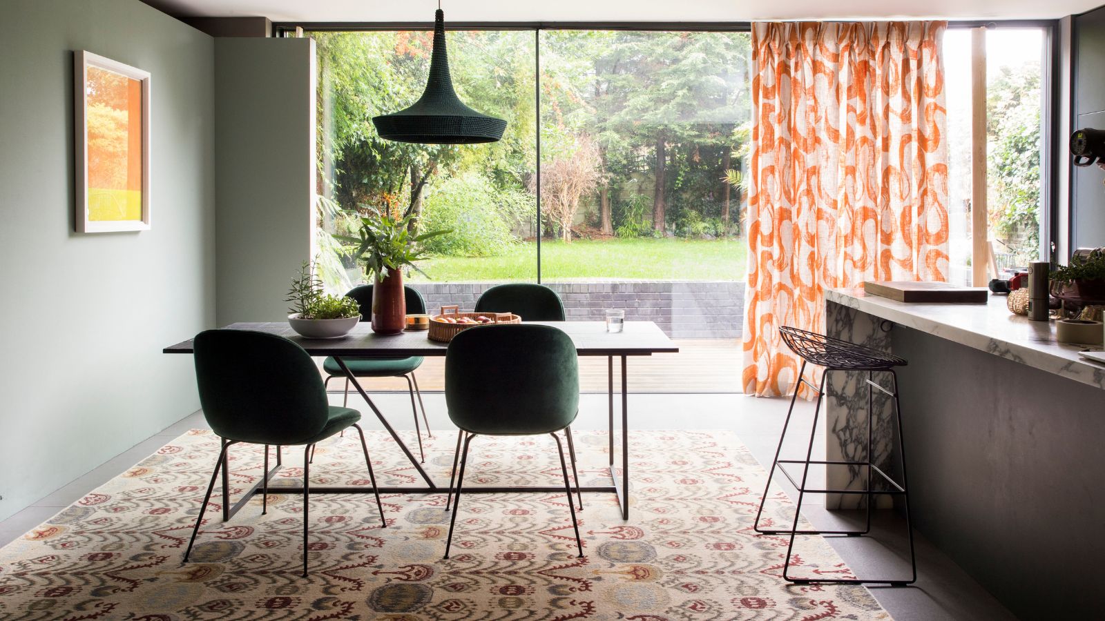 A naturally lit kitchen dining area with a black and marble island with black barstools, and a black dining table with green velvet chairs on top of a large patterned rug. Seen behind are floor to ceiling windows with an orange and white curtain, and a large green garden.