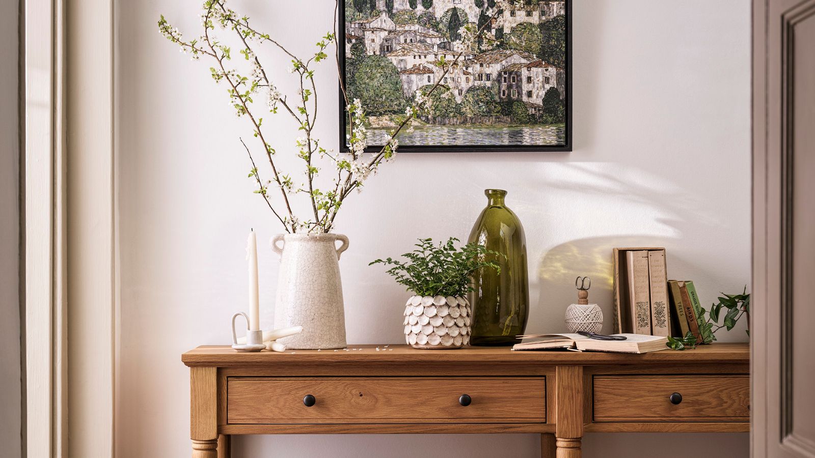 A long wooden entryway table in a cream painted entryway. A cream vase with green stems in on top, next to a small green fern in a white pot, and an empty green glass vase.