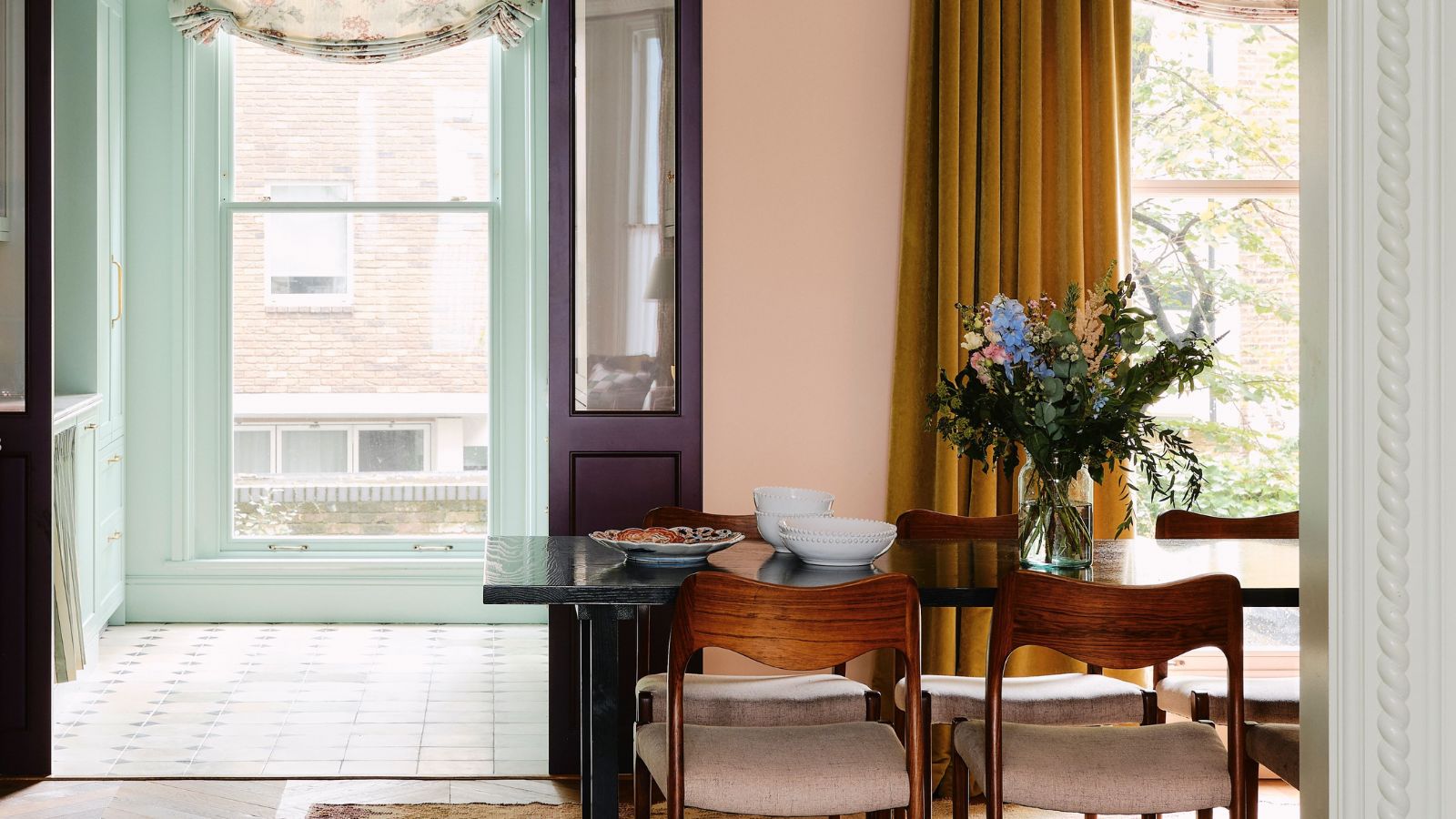 Ein pastellrosa Esszimmer mit schwarzem Esstisch und dunklen Holzstühlen. Große Türen öffnen sich, um einen pastellblauen Raum dahinter zu zeigen. 