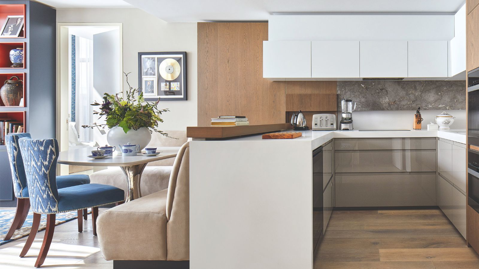 A bright kitchen with white cabinets and wooden flooring. To the left is a comfortable seating/ dinign area with a beige sofa chair, table, and blue patterned seats with wooden legs. To the far left is a blue bookshelf.