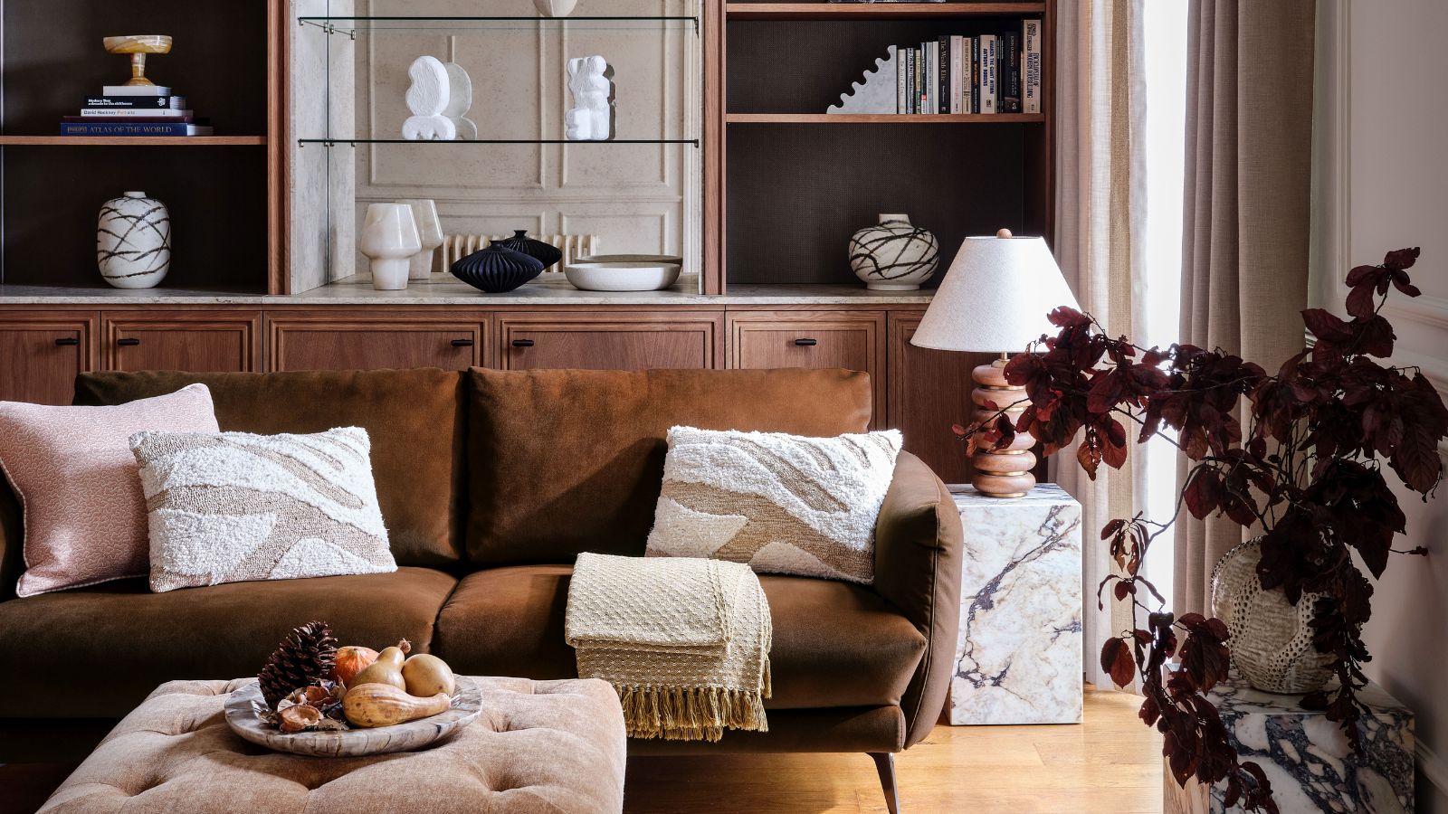 A brown velvet sofa in a neutral living room. Large wooden built-in shelves behind, a small marble table beside it, and a brown-leaf plant in the foreground.