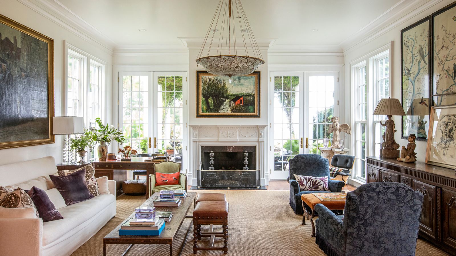 Living room with high ceilings, large fireplace with painting above, antique glass pendant, antique books and vases and upholstered armchairs