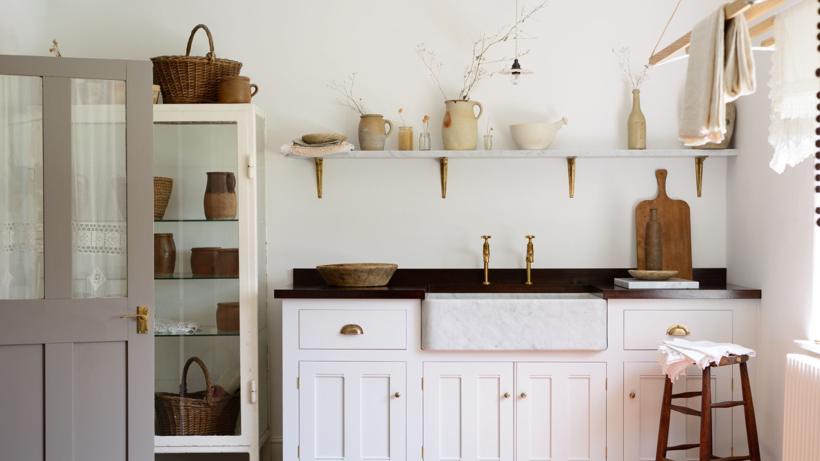 White cottage style laundry room with shaker cabinets, marble sink, brass fixtures, glass cabinet with woven baskets and natural material storage pots. There is a shelf above the sink, and a line drying apparatus mounted to the ceiling on the right