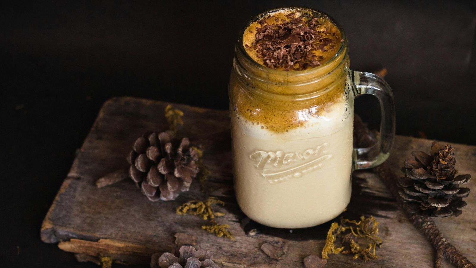 Mason jar filled with coffee on a wooden bench scattered with pinecones