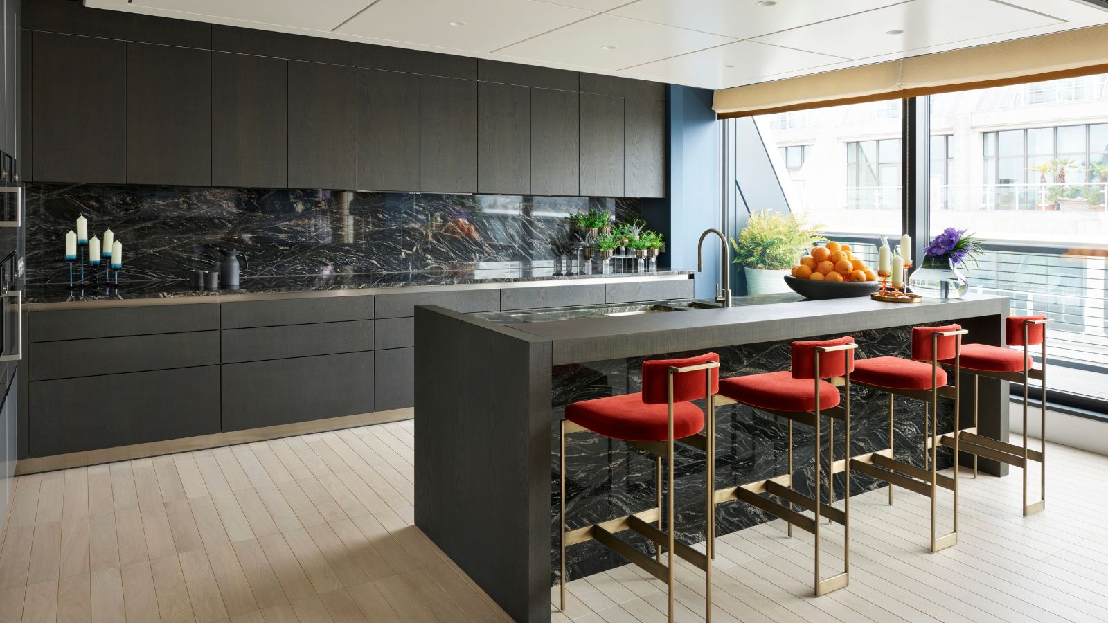 Kitchen with pale wood flooring, dark grey kitchen cupboards, kitchen island with black and white marbled worktop and splash backs. Metal bar stool with upholstered orange seats, floor to ceiling windows onto the balcony.