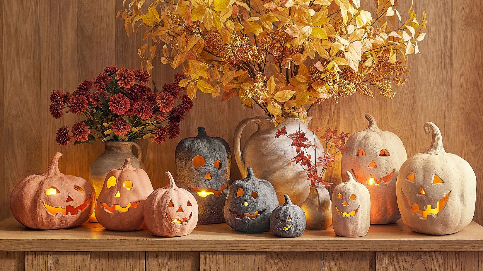 Wooden shelf displaying cream, black and orange terracotta pumpkins, alongside two large vases of red flowers and yellow leaves