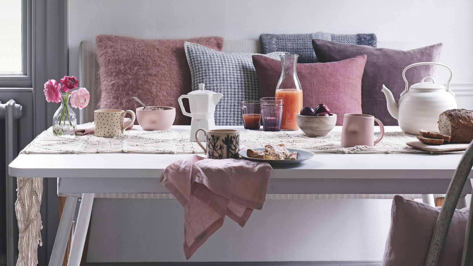 White breakfast table with pale pink napkins, pale pink dinnerware, white Moka pot and white teapot, with colorful cushions on a bench behind