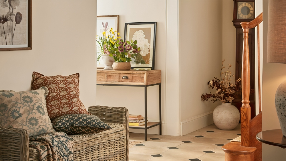 A large entryway with traditional tile flooring and a wicker storage bench with throw cushions on it. The bottom corner of a wooden stair rail visible. Through an archway, an entryway table with flowers and leaning frames on top.