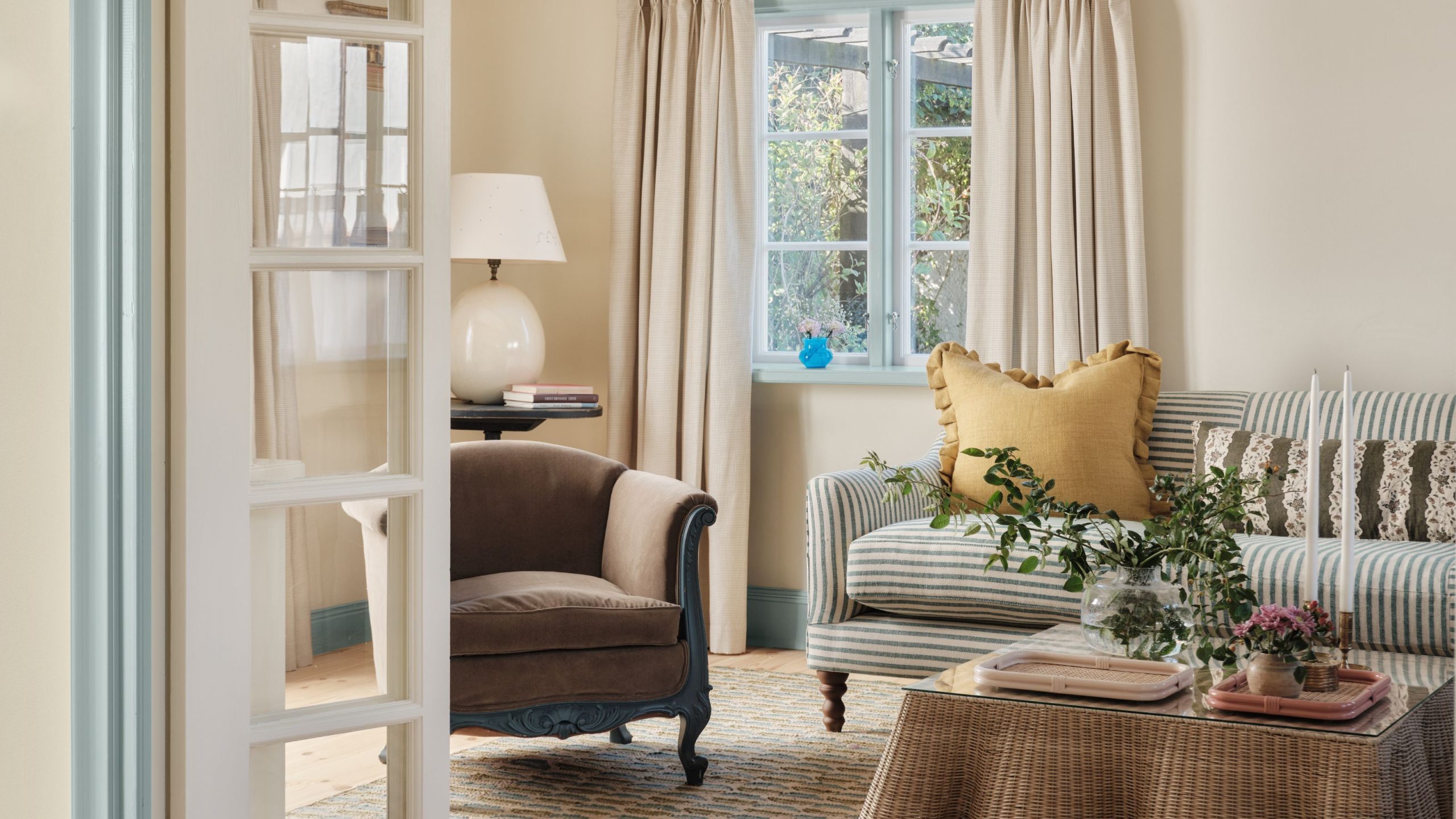 Cream living room with the windows painted in a pale blue. A striped blue and white sofa is against the far wall and a brown vintage armchair sits in front with a rattan coffee table in the center