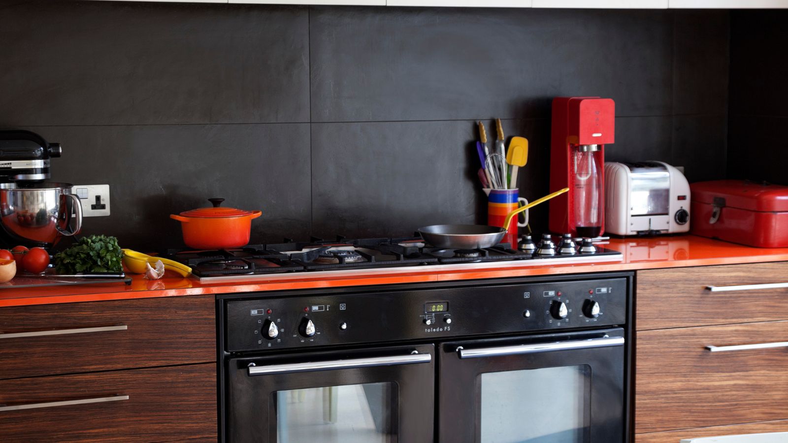 Kitchen with dark wooden cabinets, silver handles, black double oven, red counter tops and red appliances