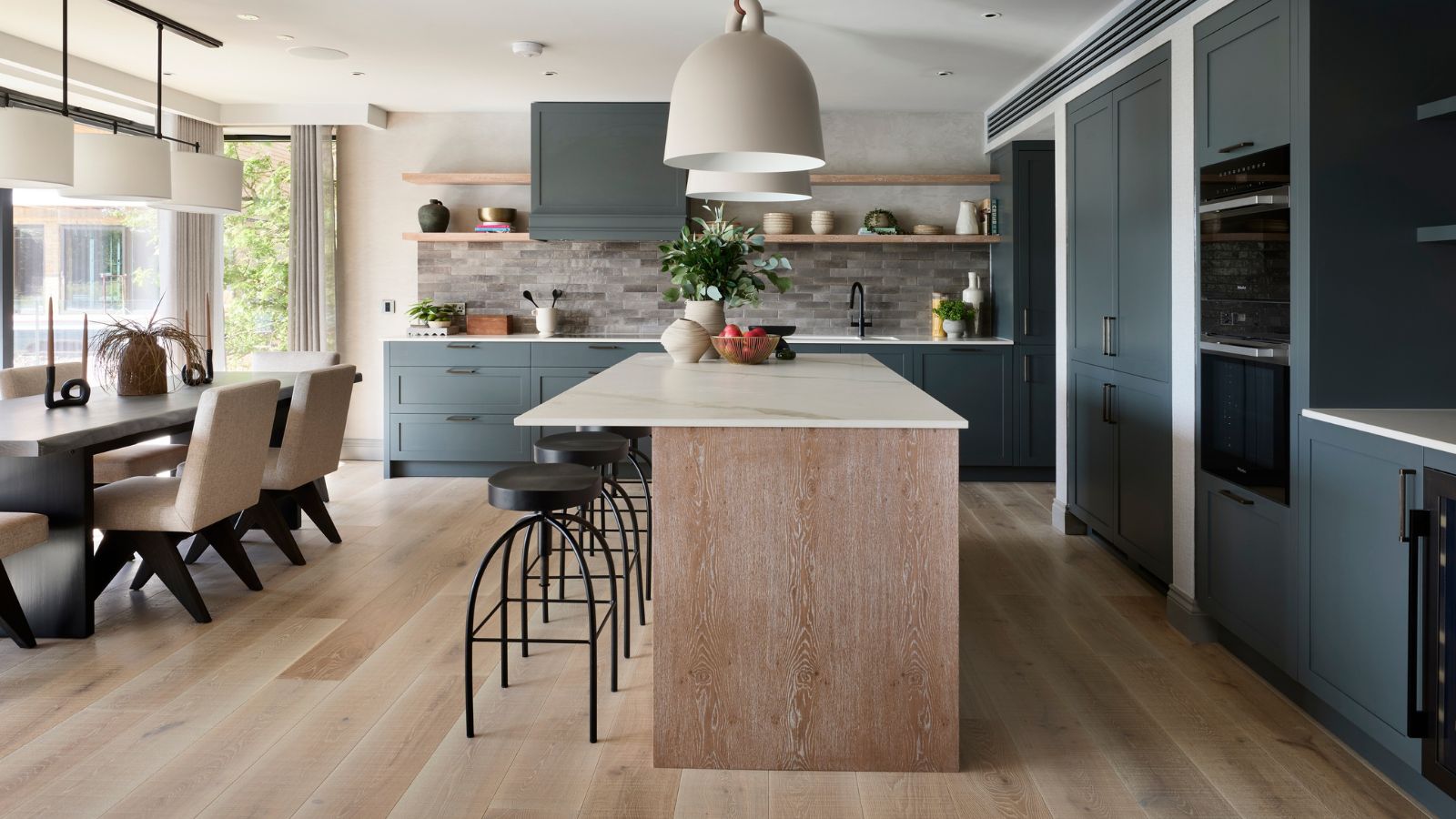 A dark blue fitted kitchen diner, with a large wooden island. Industrial style black barstools pulled up to the island, with a large black dining table behind it, with white upholstered chairs.