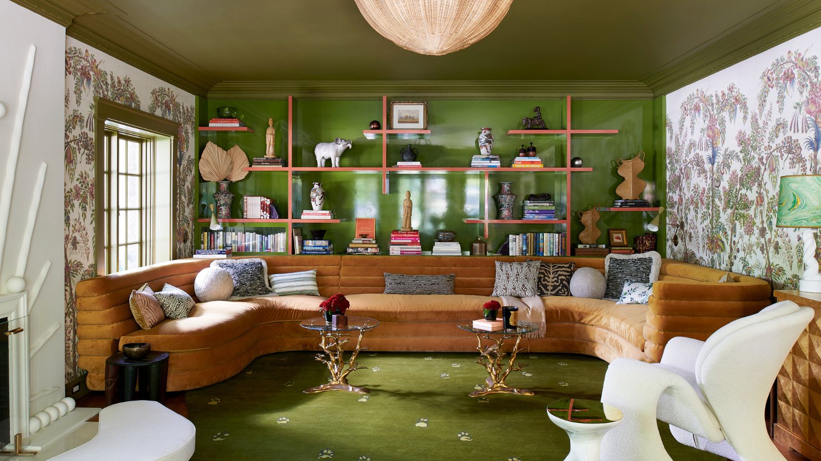 Living room with green painted ceiling and wall, open shelving, wallpaper mural, green rug and large U-shaped brown sofa with modern armchair and two ornate iron side tables