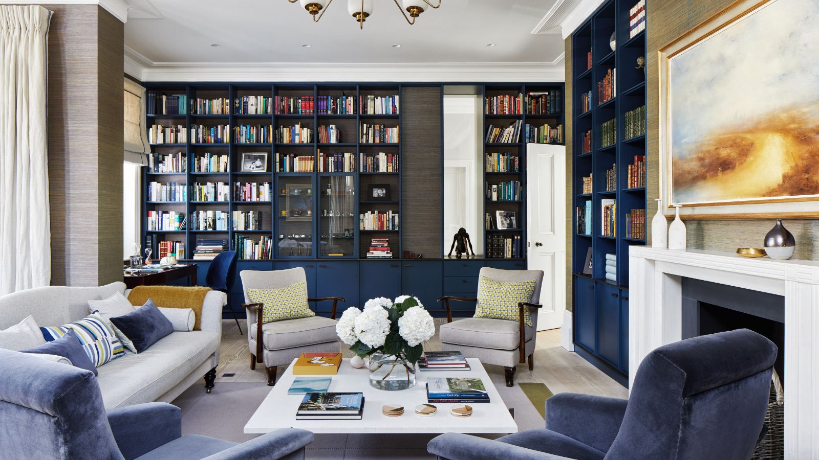 Living room with gray walls, dark blue fitted bookcase and display units, grey and blue sofa and armchairs around a white coffee table beside the fireplace.