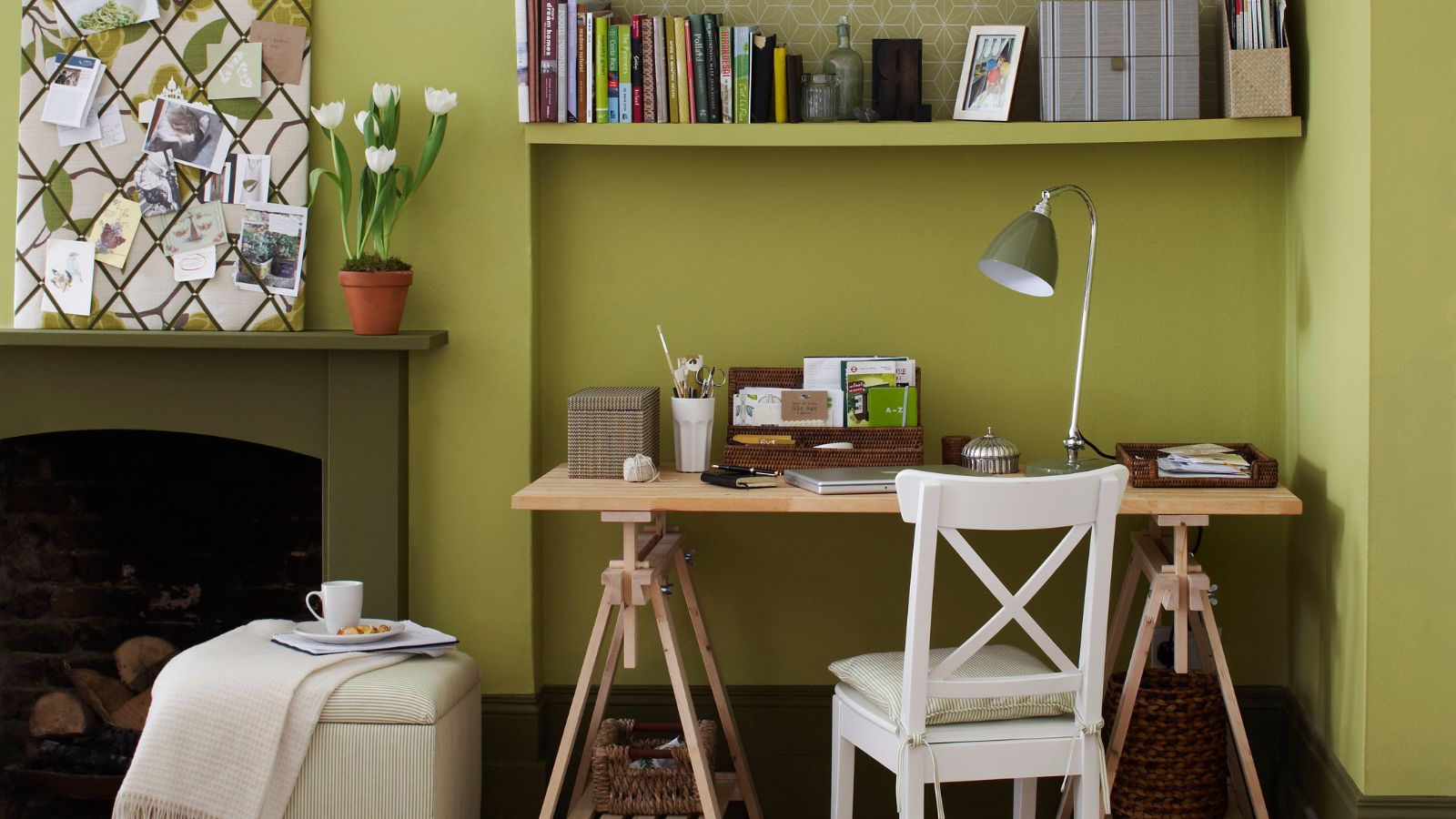 Home office in alcove of green living room, patted storage ottoman, desk, white chair, shelves in alcove, fireplace, fabric covered notice board.