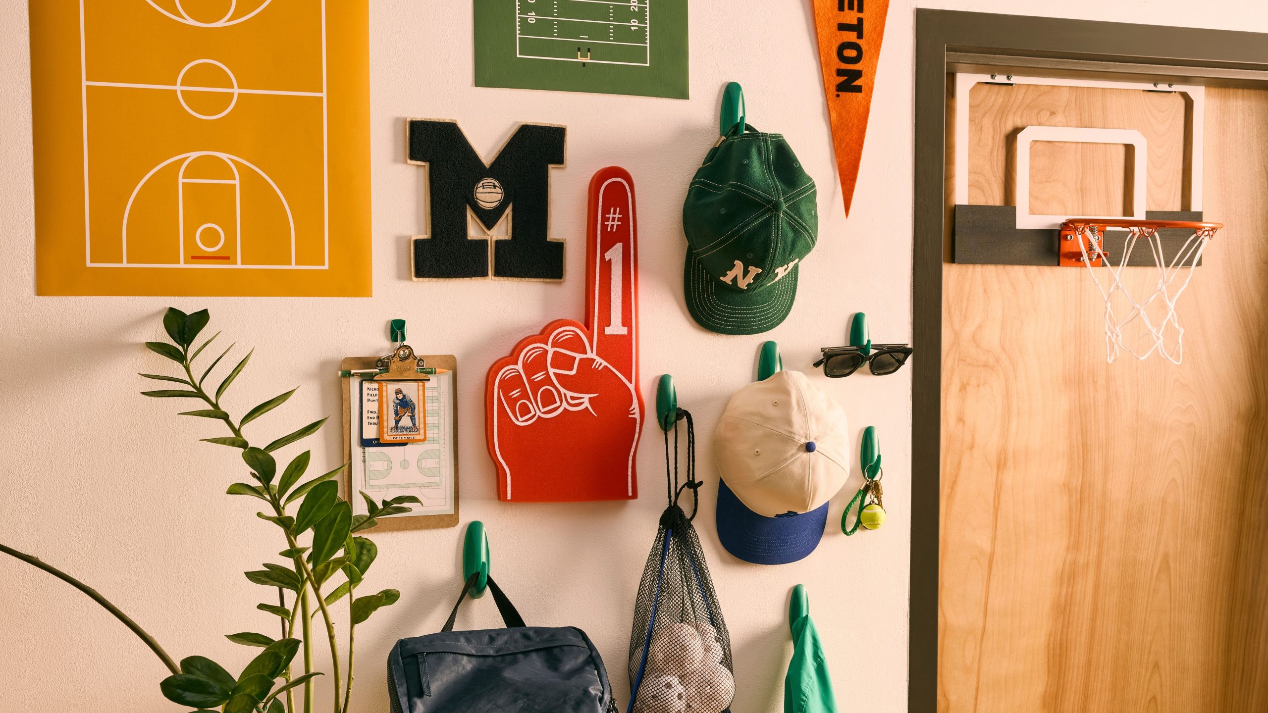 Dorm room decor, including a foam finger, collegiate monogram letter, ball caps, and posters, mounted to a wall alongside a mini basketball hoop, which is mounted to the door