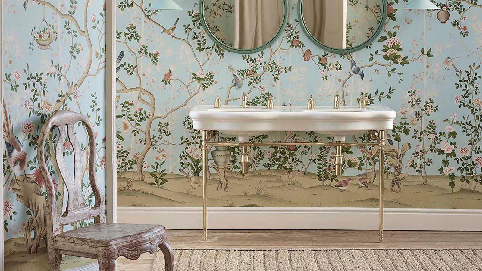 Bright, large bathroom with pale blue scene wallpaper feature and Victorian double sink with gold hardware, and a vintage, distressed chair