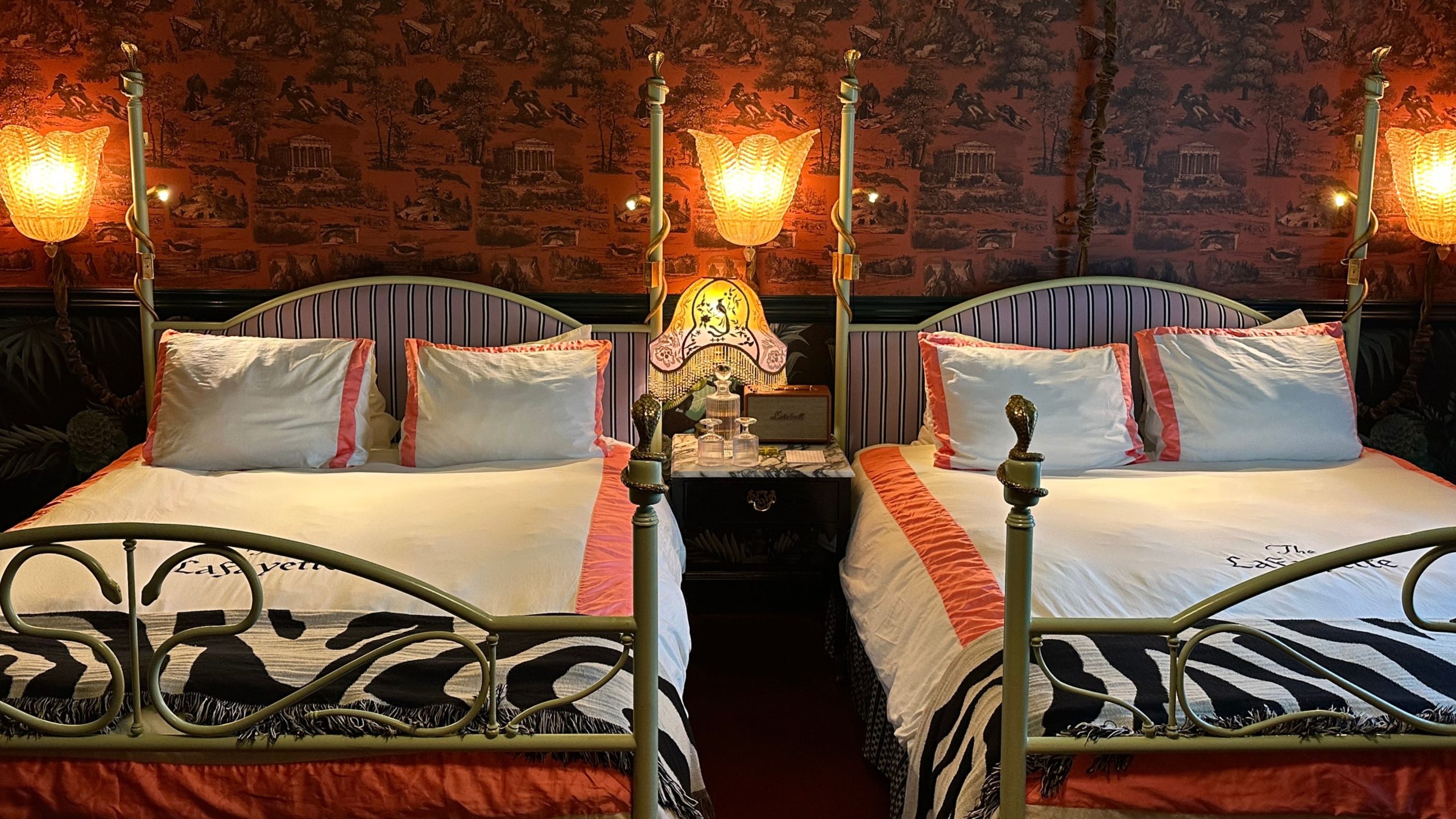 Eclectic guest room at the Lafayette Hotel in San Diego, featuring two four-post beds with striped headboards and pink bordered bedding, along with pink toile wallpaper and a fringe antique-style table lamp.
