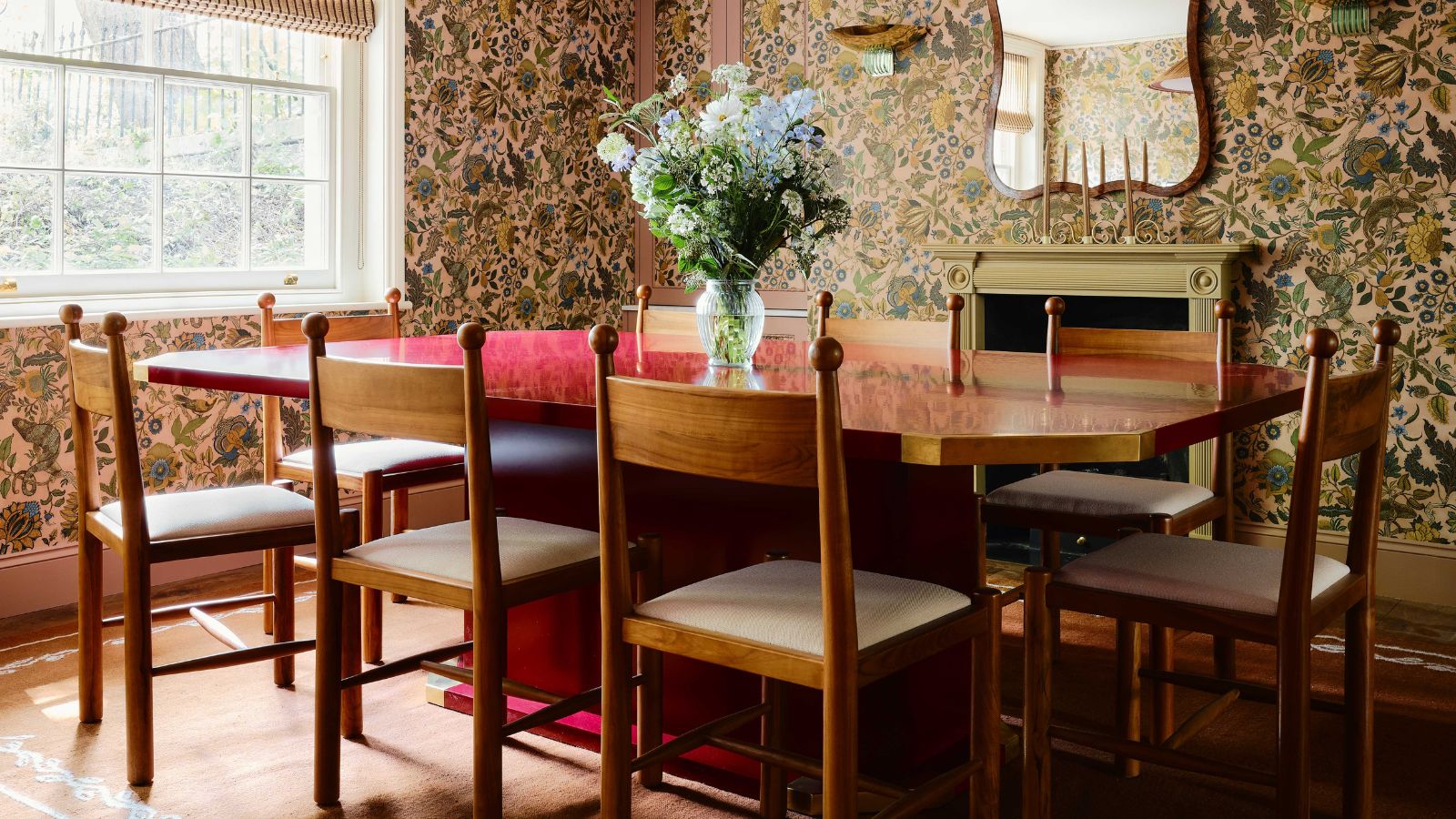 large dining room with beige and green/blue floral wallpaper with a fireplace and large window with a large red lacquer dining table and retro pine chairs