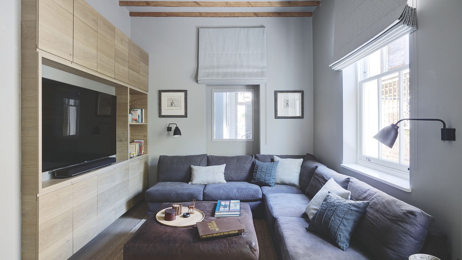 A cozy living room with a large L-shaped sofa, grey cushions, a leather ottoman and large television in a wooden shelving unit. The walls are white with artwork and lamps, and the windows have pale grey open blinds on them. There are exposed beams on the ceiling.