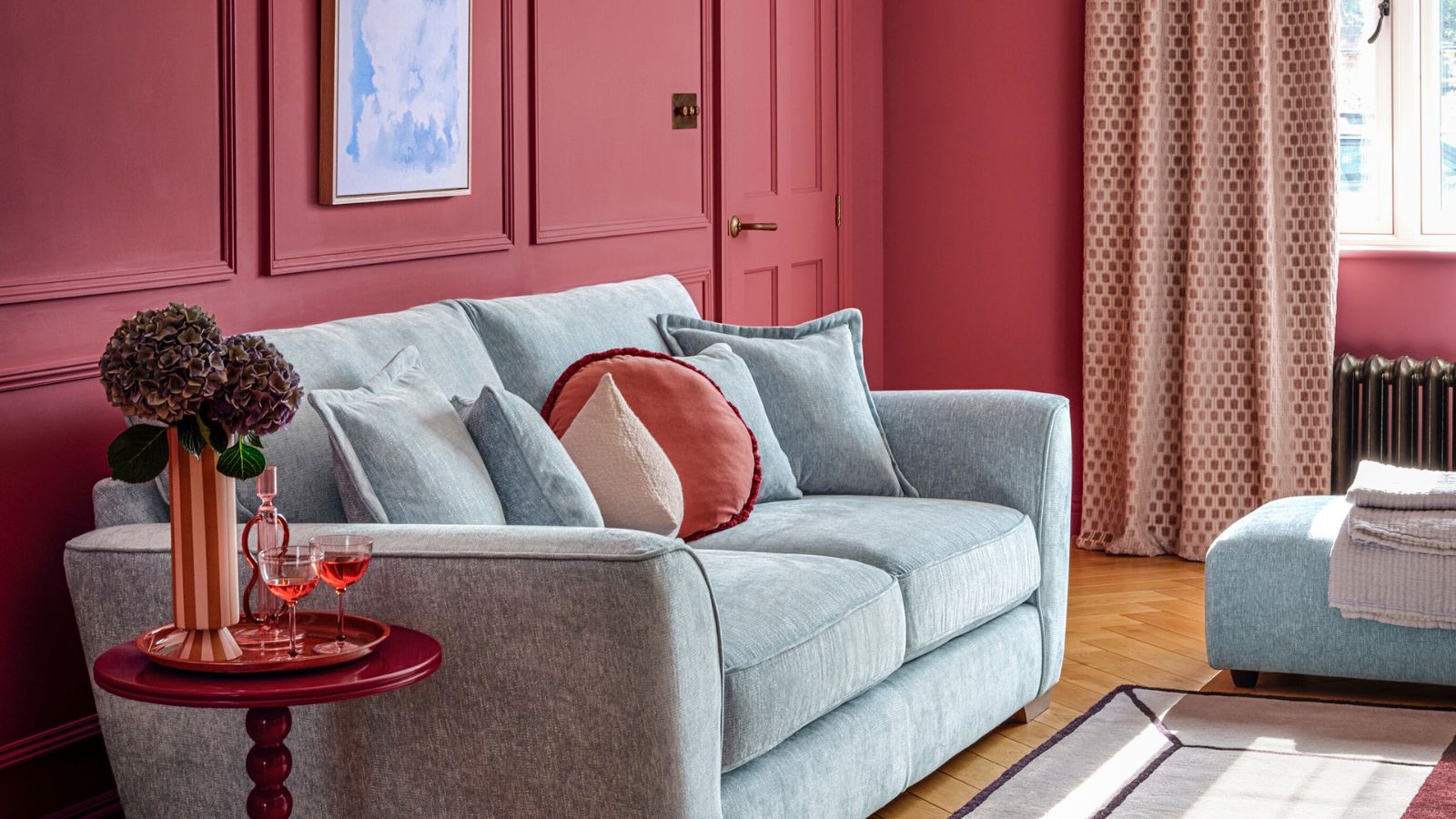 A red painted living room with a light blue sofa, with red cushions on it. A small red side table beside the couch with a tall vase of flowers on it.