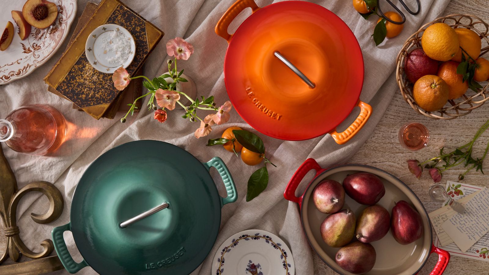 A green and orange Dutch oven from the new Le Creuset modern heritage collection, shot from above, surrounded by fresh fruit and flowers