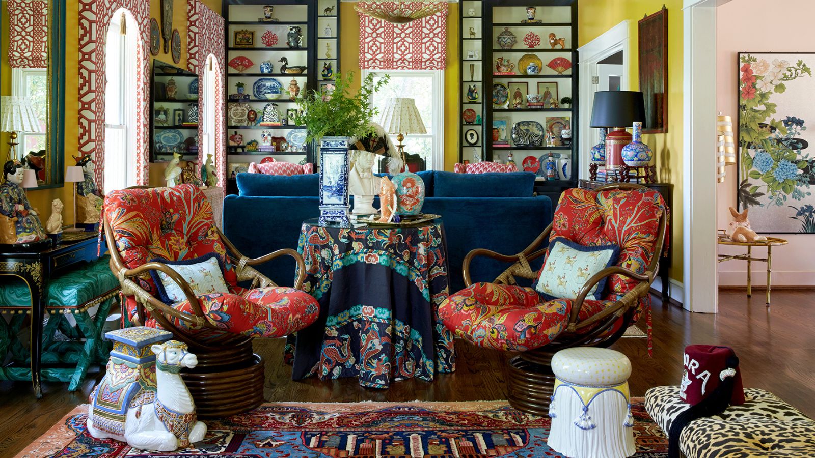 maximalist living room with yellow walls, bookcases filled with antiques, large blue couch, red upholstered antique chairs, ceramic stool and leopard bench