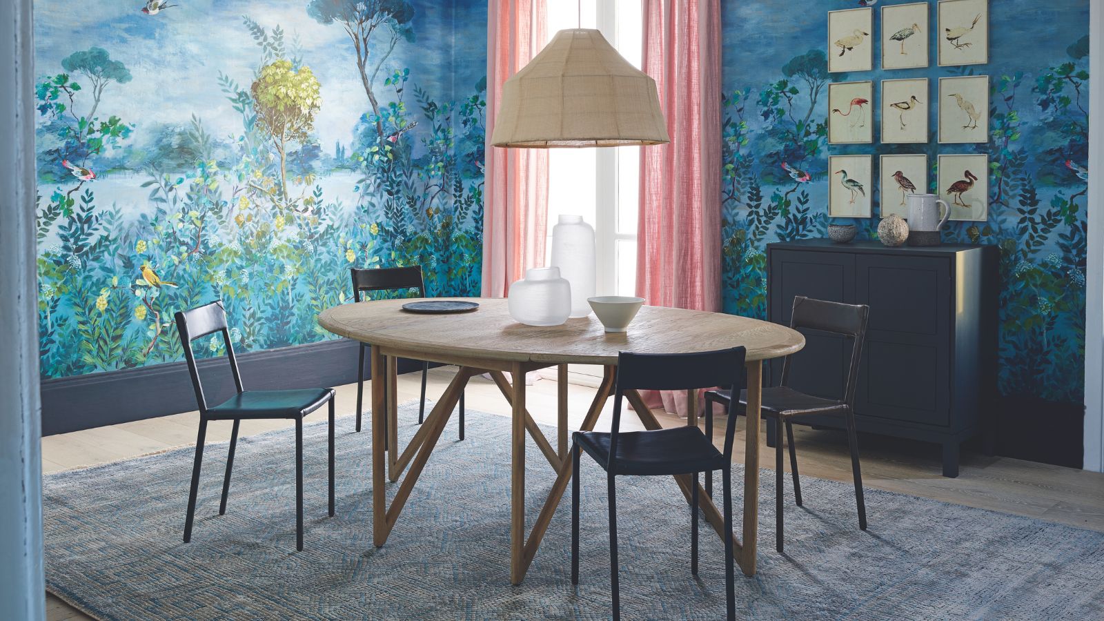 A blue dining room with patterned statement wallpaper, pink curtains, a rattan lamp, round wooden table and black chairs.
