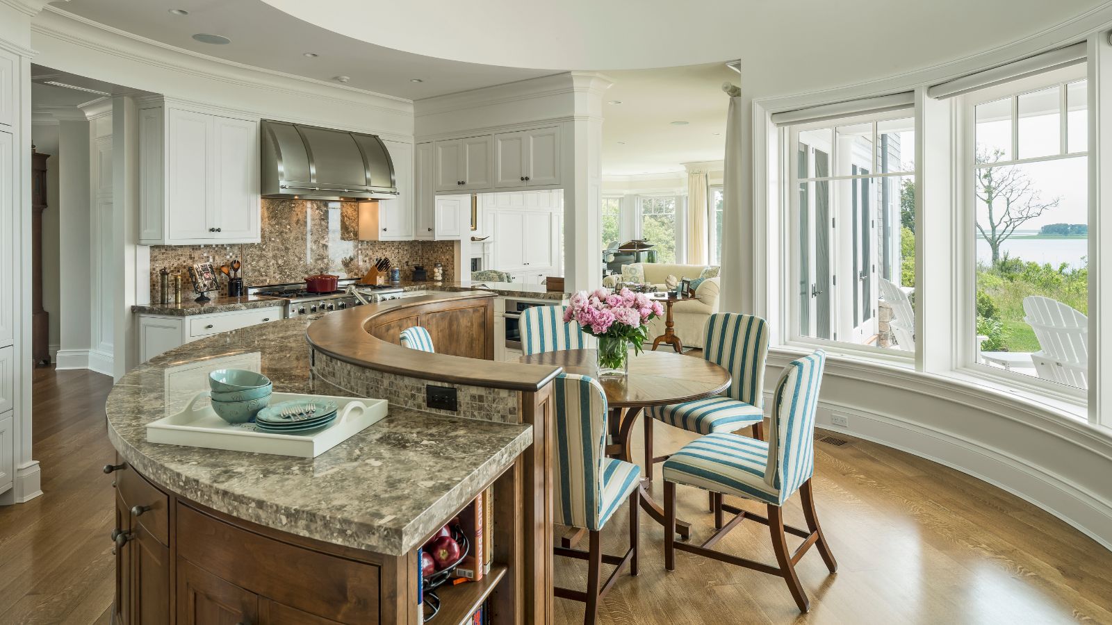 Bright coastal home with rounded kitchen features and layout. There is a marble island in a semi circle, striped blue and white upholstered chairs around a round table and bright tall windows with the ocean in view