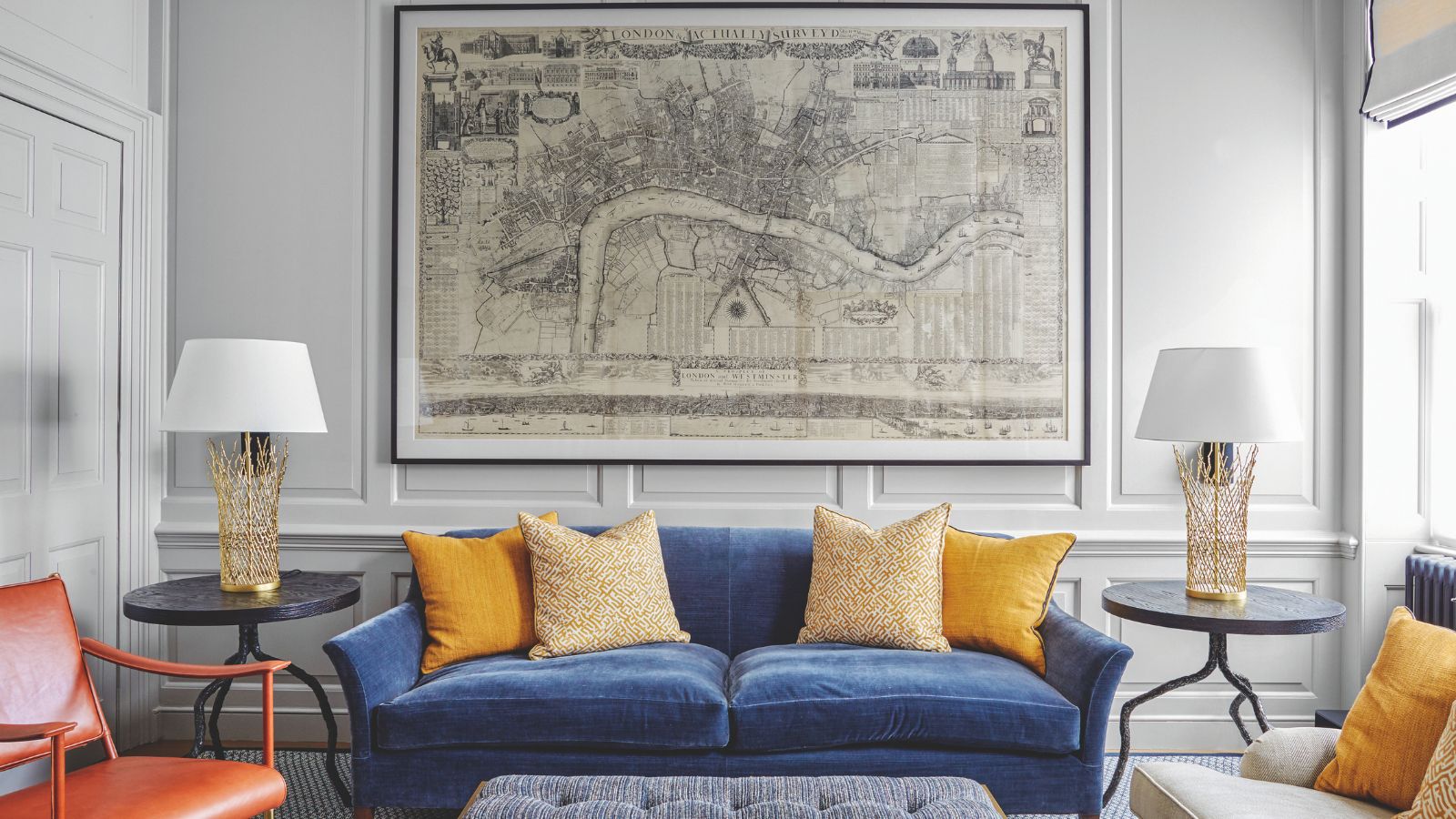 A white living room with panelled walls, a blue velvet sofa with yellow cushions, an orange chair, padded upholstered ottoman, two matching statement gold lamps with white lampshades, and a large map artwork on the wall.